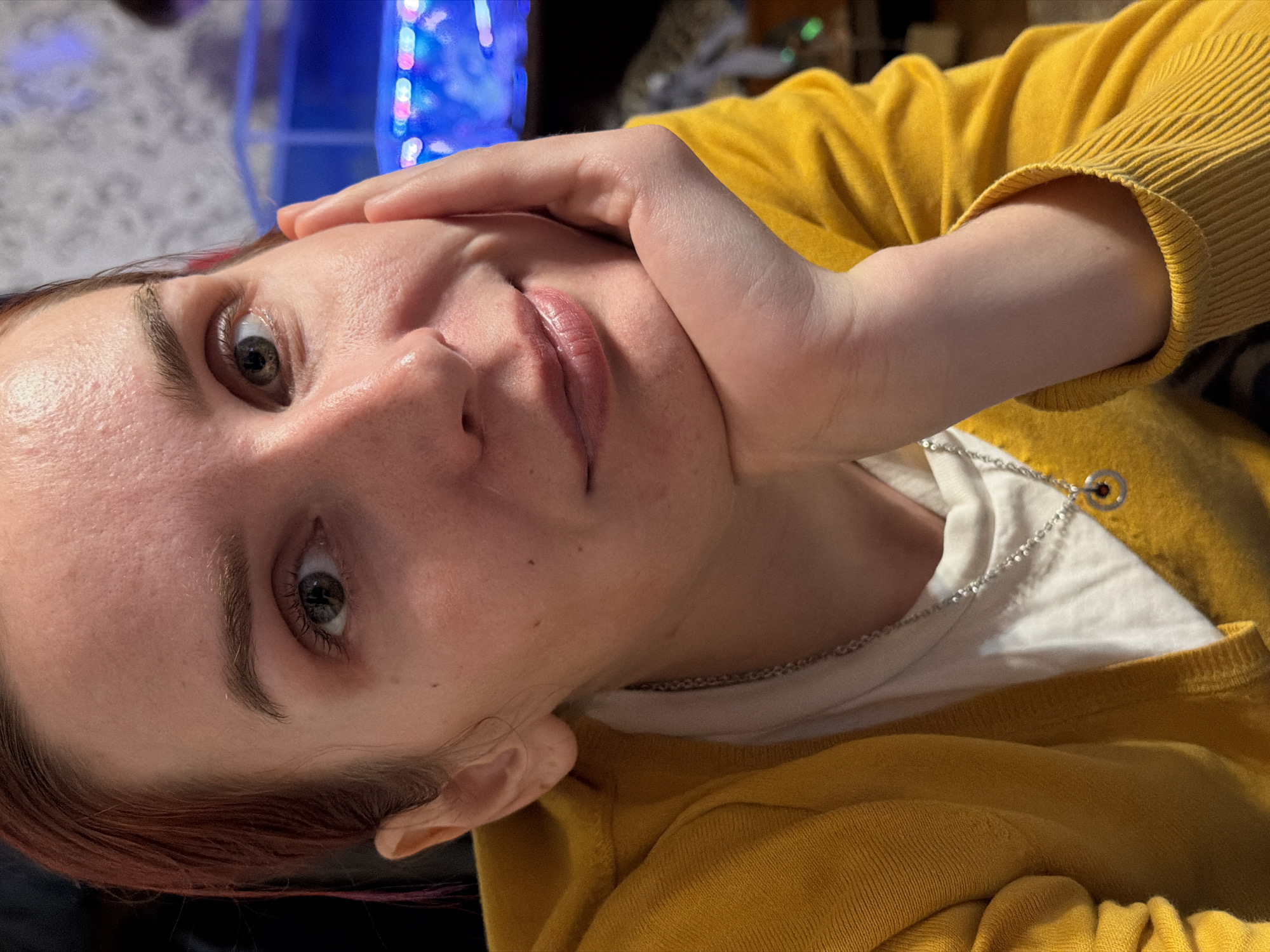 This is an angled, close-up portrait of a person, likely a woman, set indoors in what appears to be a home in Заболотово, Russia. The subject is positioned diagonally across the frame, looking directly at the camera.
The primary subject is a woman with fair skin, light-colored eyes (appearing hazel or grayish-green), and reddish-brown hair. Her eyebrows are dark and distinct. Her lips are a natural pink color, and her skin shows natural texture with some minor blemishes. She has a neutral to slightly pensive expression, with her eyes open and engaged.
She is wearing a mustard yellow, ribbed knit cardigan over a plain white t-shirt. A thin silver chain necklace with a small, simple circular pendant hangs around her neck. Her left hand is resting against her face, with her chin cradled, suggesting a relaxed pose or a moment of contemplation.
The background is out of focus but reveals blurs of bright, colorful artificial lights, possibly from an electronic device, an aquarium, or decorative lighting, consistent with a domestic setting. The image lacks specific cues for the time of day, but the artificial lighting in the background hints at evening or low light conditions indoors. There is no visible text in the media file.