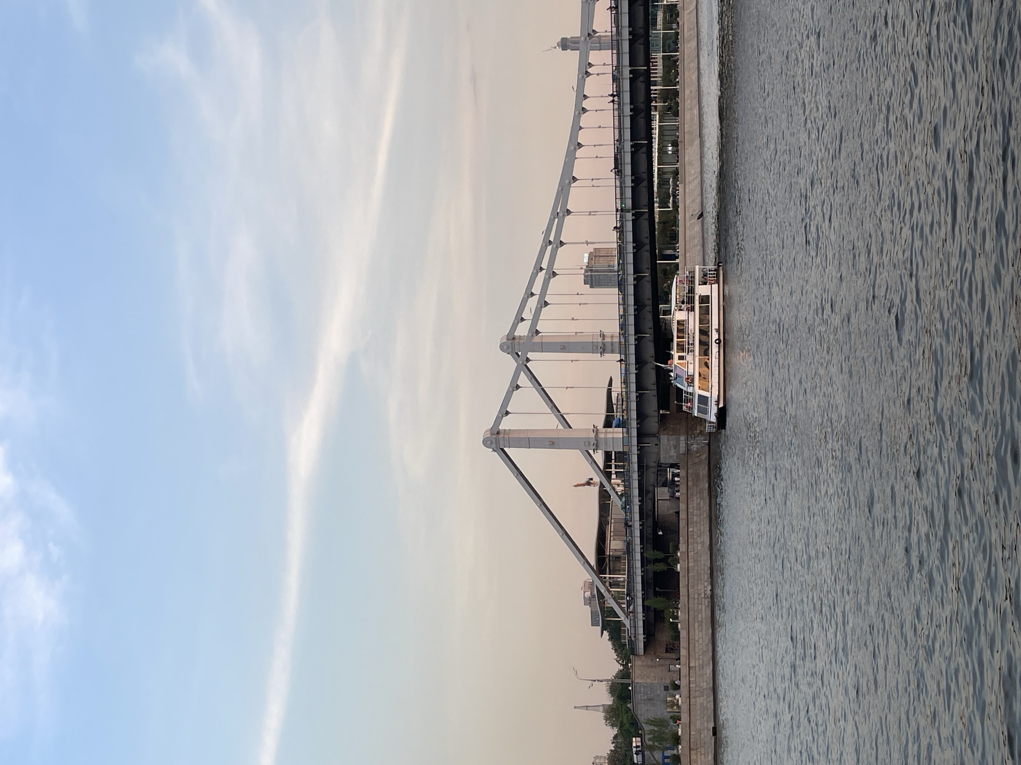 The image shows a bridge over a river in Moscow, Russia. A boat is docked next to the bridge. The sky is light blue with some wispy clouds. The water is gray and choppy. The bridge is gray and has a series of arches. There are buildings and trees on the riverbank.
