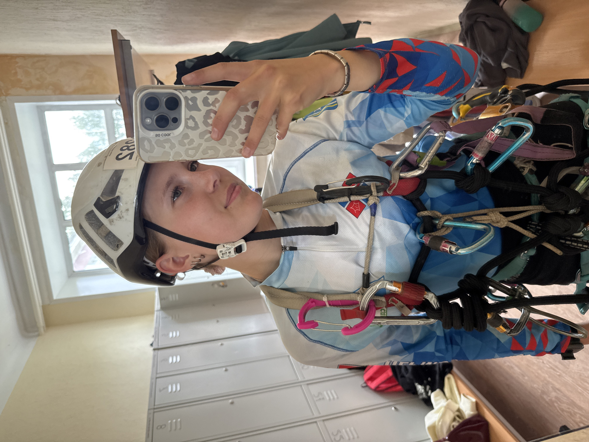 The image shows a young person in what appears to be a locker room, possibly before or after a sports activity. The person is wearing a white helmet with some stickers on it, and a brightly colored long-sleeved shirt with a geometric pattern of red, blue, and white. They are also wearing a climbing harness with multiple carabiners and ropes attached, suggesting they are involved in climbing or a similar activity. The person is holding up a smartphone in a leopard-print case to take a selfie in a mirror, reflected in which is the locker room.
Visible in the background are rows of grey metal lockers, a window with a view of trees outside, and a wooden cabinet. The lighting suggests it is daytime. The person's facial expression is neutral and focused on taking the photo. The overall scene is casual and practical, related to sports or outdoor activities.
The stickers on the helmet are partially visible, with one sticker showing "20 COOR". The phone case has a leopard print pattern in black and white, and the camera lenses are clearly visible. The harness and climbing equipment are detailed, with various carabiners in different colors like silver, blue, and pink, and black ropes.
The setting appears to be a functional space designed for sports participants, likely in Chaikovsky, Russia, given the provided location context.