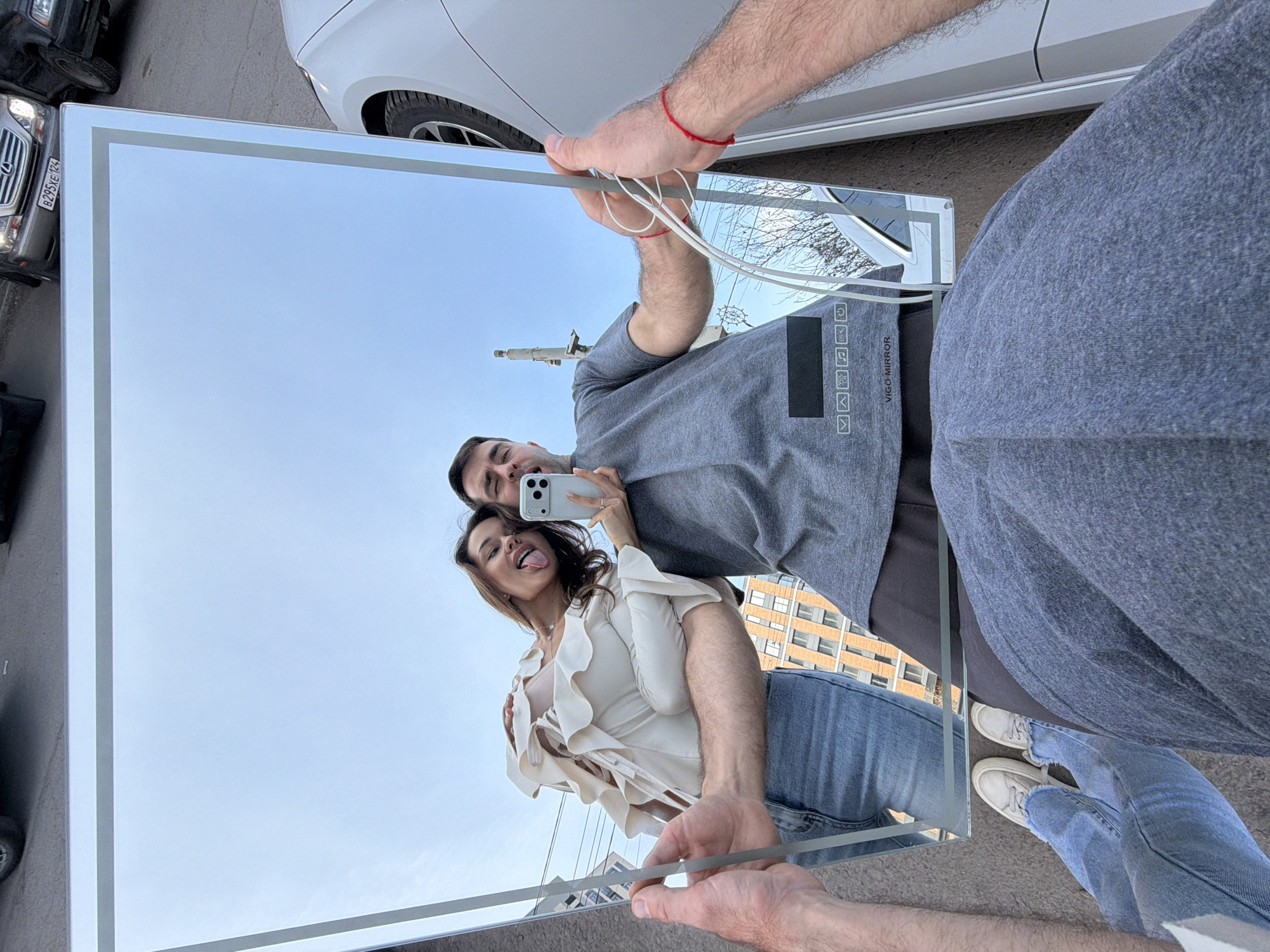 A unique, rotated photograph captures a couple taking a selfie using a large, rectangular mirror held by the man, reflecting their images against an outdoor urban backdrop. The image is oriented 90 degrees clockwise, making the ground appear on the left.
In the foreground, the man's arm, adorned with a red string on his wrist, holds the mirror. His reflection in the mirror shows him holding a white smartphone, seemingly taking the picture, with a gentle smile. Beside him in the reflection, a woman playfully sticks out her tongue, conveying a joyful and lighthearted mood.
The woman is dressed in a light-colored, ruffled blouse, blue jeans, and white sneakers, while the man wears a grey t-shirt and grey sweatpants. The mirror itself appears to have an illuminated border. Within the mirror's reflection, behind the couple, several urban buildings are visible under a clear, bright blue sky, indicating daytime with good weather.
Outside the mirror's reflection, parts of two cars are visible: a white sedan and a dark SUV with a Russian license plate "B295YE 124," confirming the location context of Krasnoyarsk Krai, Russia. The man's grey t-shirt, seen in the mirror's reflection, features the vertical text "VIGO MIRROR" along with various icons including a checkbox, up arrow, refresh symbol, play/pause button, music note, Wi-Fi symbol, and power button, suggesting potential smart mirror functionalities.