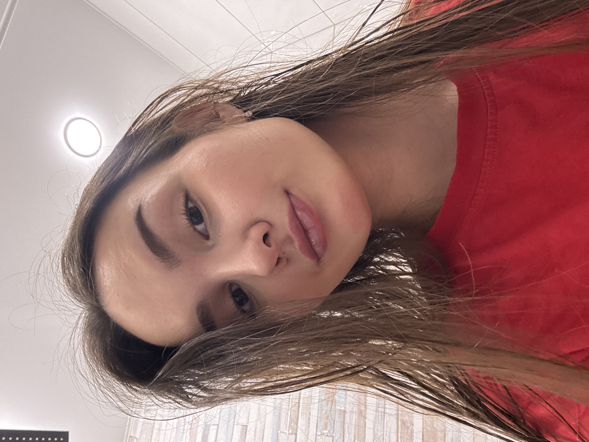 A young woman with long, straight brown hair is the primary subject of the image. She is wearing a red shirt. Her face is turned towards the camera, and she appears to be looking directly at the viewer. The background includes a tiled wall and a ceiling with a recessed light fixture. There is no evidence to confirm that the location is in the city of Chaikovsky, Russia. The description is limited to the visual details present in the image.