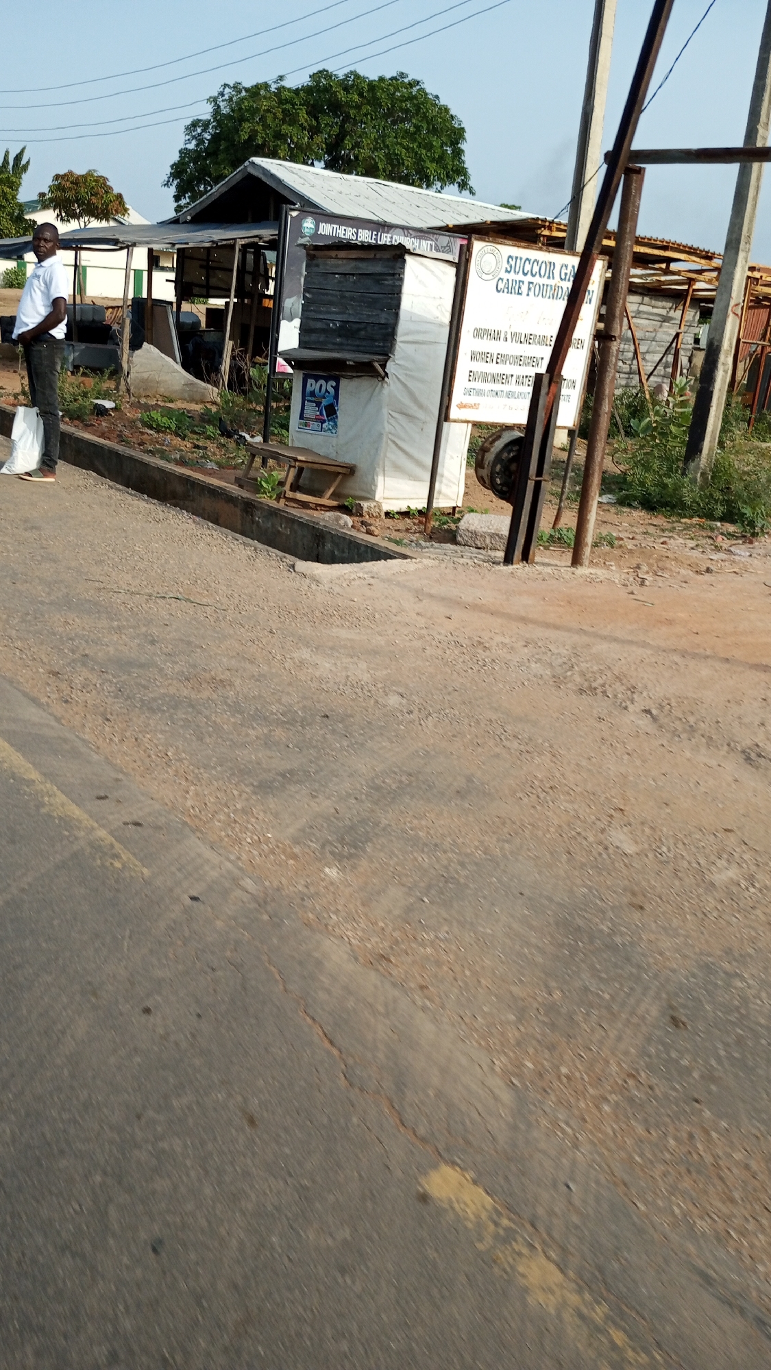 A scene in Otokiti, Nigeria shows a man in a white shirt and dark pants standing next to a concrete curb, holding a white plastic bag. Behind him is a small, rudimentary structure with a sign reading "JOINTHEIRS BIBLE LIFE CHURCH INT'L" and a "POS" advertisement. Adjacent to this structure is a larger sign for "SUCCOR GA CARE FOUNDAN," listing services like "ORPHAN & VULNERABLE REN," "WOMEN EMPOWERMENT," and "ENVIRONMENT WATER." The sign also mentions "SHETHIRA OTOKITI NEW LAYOUT." The foreground shows an asphalt road with cracks and yellow paint. A utility pole stands to the right of the signs. Vegetation and other makeshift structures are visible in the background.