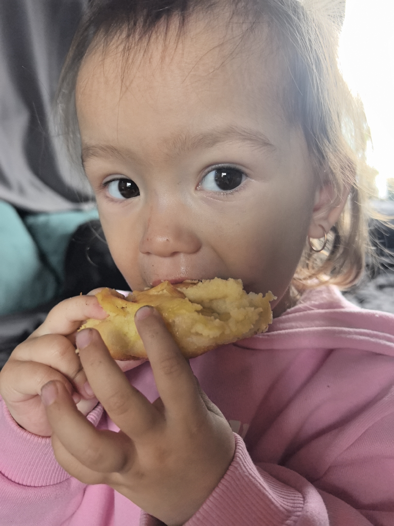 A close-up portrait features a young child, likely a toddler, actively eating. The child has dark eyes and light brown hair, wearing a pink hooded sweatshirt. A small, thin gold hoop earring is visible in the child's right ear. The child is holding a piece of yellowish food, which appears to be a pastry or a baked item, possibly containing potato, and is in the process of taking a bite. Traces of food are noticeable around the child's mouth and nose, suggesting a messy but enjoyable snack.
The child looks directly into the camera with a focused expression. The background is softly blurred, revealing hints of an indoor setting with what appears to be a grey and teal/turquoise upholstered object, possibly a couch or pillow. Bright natural light, likely from a window, illuminates the scene from the right side, creating highlights on the child's hair and side of the face, indicating it is daytime. There is no visible text in the image. The scene conveys a moment of childhood simple pleasure within a home environment in Noviy, Russia.