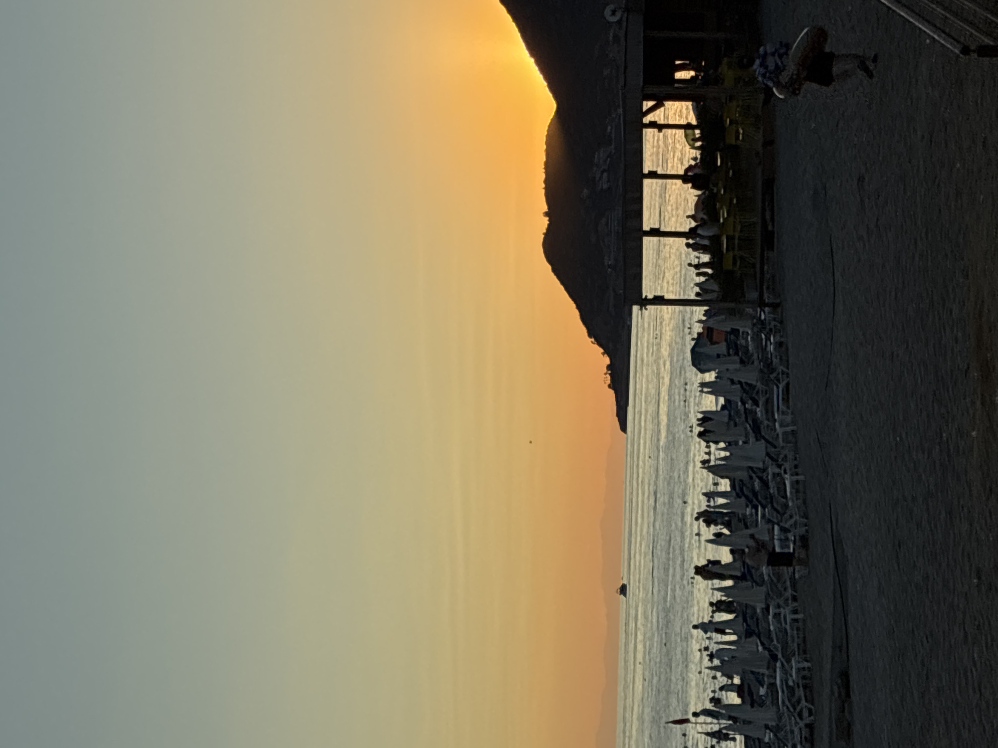 A silhouetted sunset over the sea near Alanya, Turkey, casts a warm glow on a beach lined with sun loungers and umbrellas. The sun is setting behind a dark, mountainous silhouette. In the foreground, a beach with dark sand is partially visible, with a few people walking. To the right, a shaded structure with tables and chairs suggests an outdoor seating area for a beachside establishment. In the water, a small boat is visible, and further in the distance, a hazy coastline with hills can be seen. The sky transitions from a light blue at the top to a vibrant orange and yellow near the horizon, indicating the end of the day.