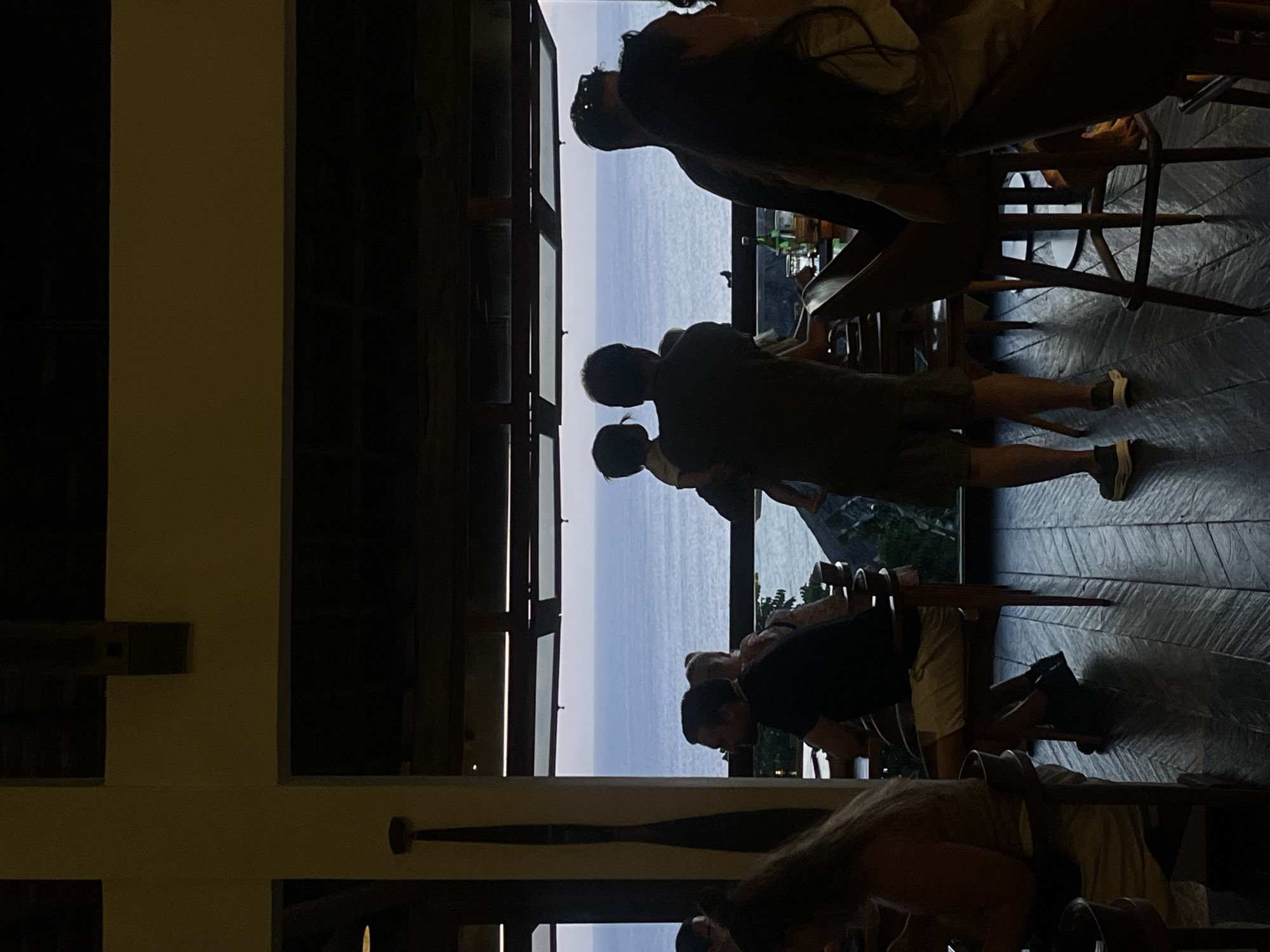 The image shows a group of people observing a coastal view from an elevated indoor space in Kuta Selatan, Indonesia. Individuals are standing and sitting, some leaning against railings or seated at tables. The background features the ocean horizon visible through architectural frames or windows. The scene is dimly lit.