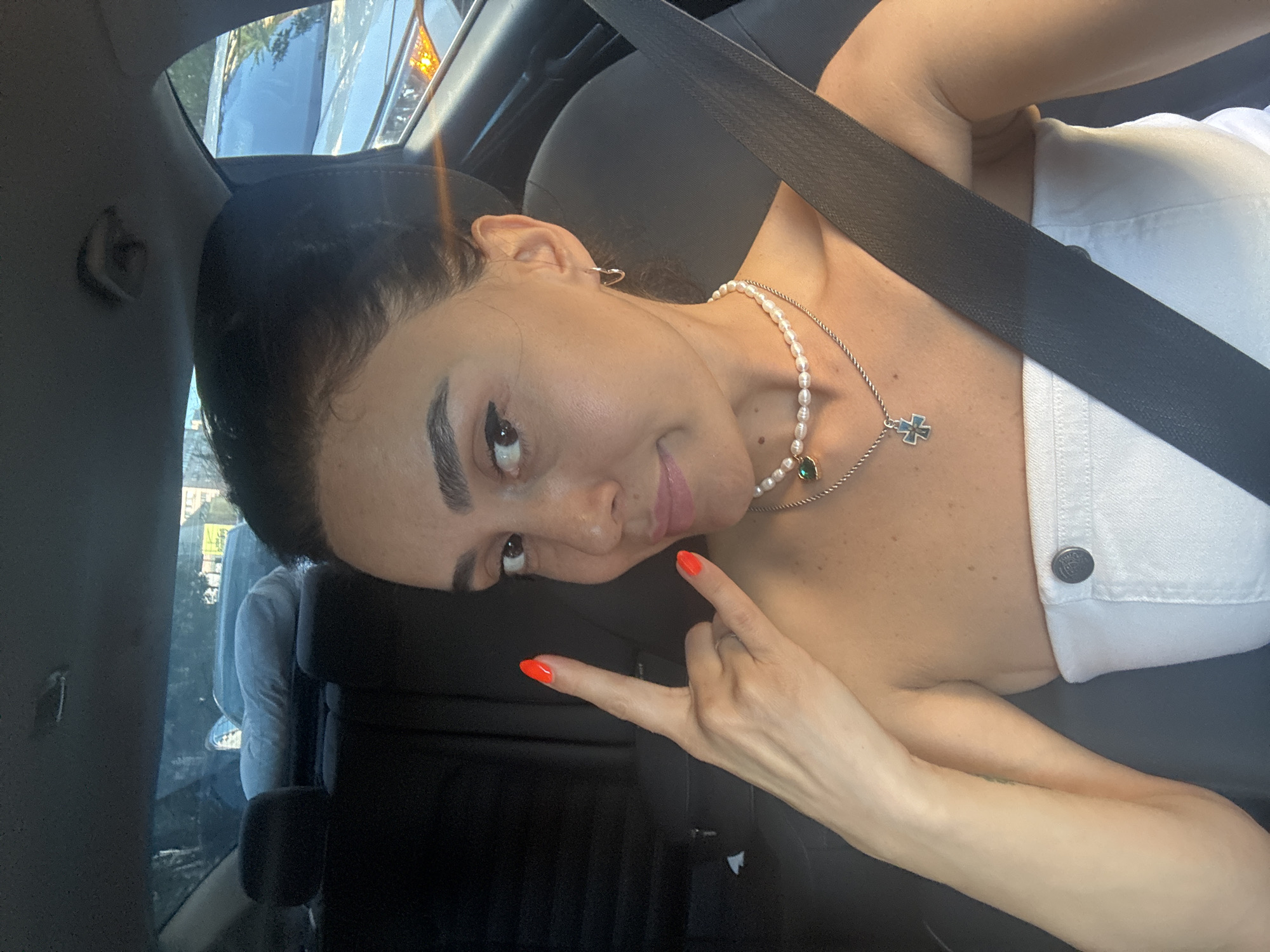 A woman with dark hair is in the passenger seat of a car, wearing a white strapless top and two necklaces. She has bright orange nail polish on her fingers and is making a "rock on" hand gesture with her left hand. A seatbelt is across her body. The interior of the car is visible, including the headrests of the back seats. Outside the car, other vehicles and some trees and buildings can be seen. The scene appears to be a street in Tbilisi, Georgia.