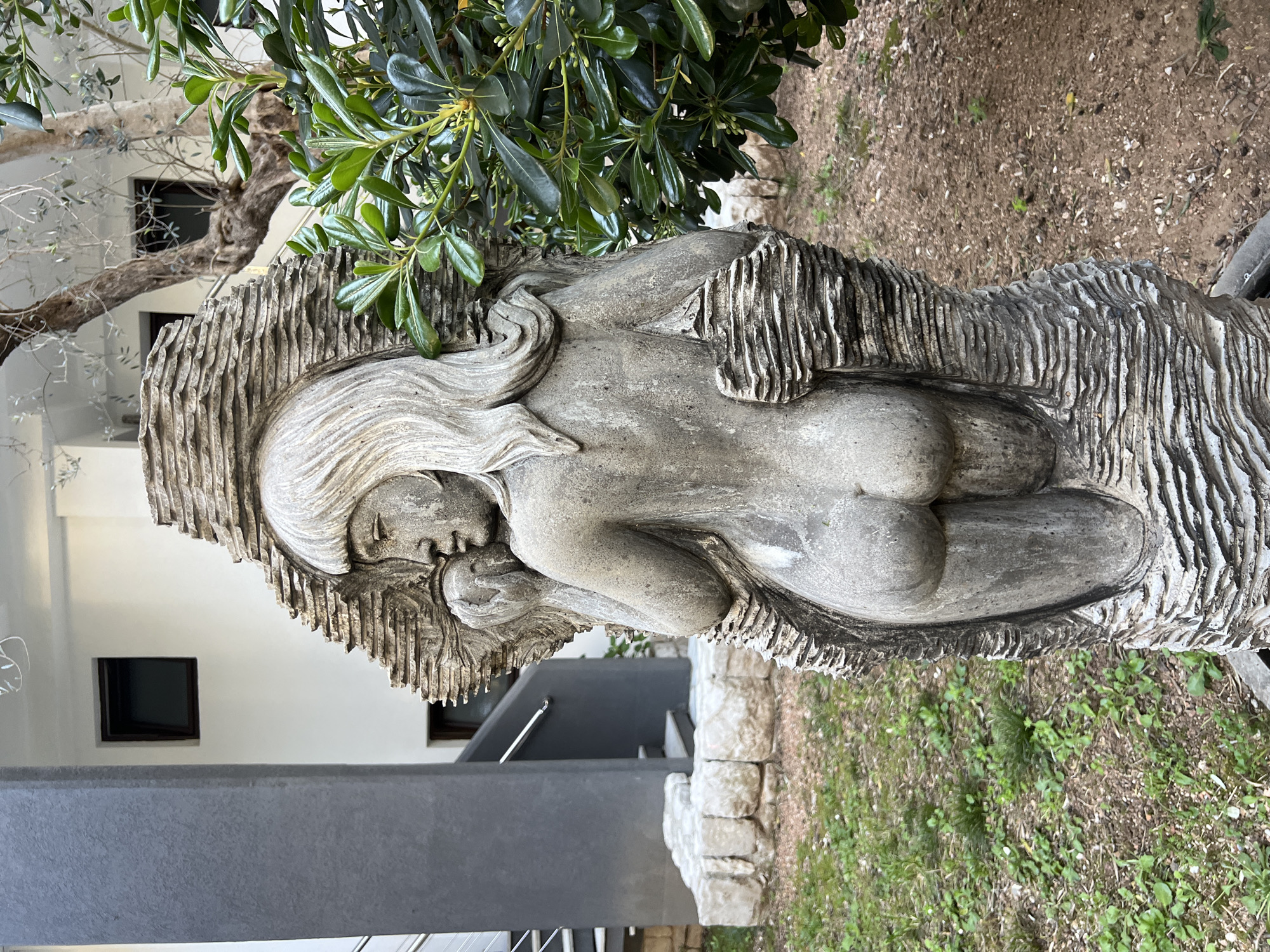 The image shows a stone sculpture of a nude woman in a garden or yard setting. The sculpture depicts a reclining female figure with long hair, her body partially integrated into a textured, stone background. Vegetation, including green leaves and branches, is visible behind the sculpture. In the background, there's a glimpse of a building with dark-framed windows and a gray architectural element, possibly part of a stairway or a balcony. The ground around the sculpture is covered with grass and dirt. While the location is suggested to be Petrovac na Moru, Montenegro, this is not evident from the image itself, as the setting could be any similar Mediterranean location.