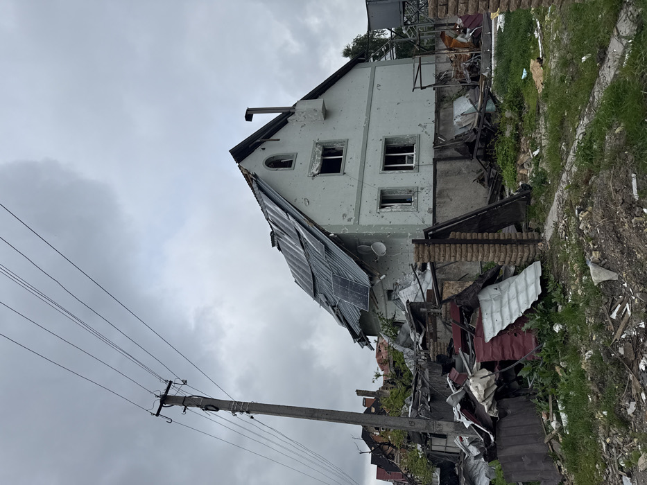 A damaged two-story house stands with holes in its walls and broken windows. Part of the roof is collapsed, displaying solar panels. Debris and rubble litter the ground around the house, signifying destruction. An electrical pole and wires are visible in the foreground against a cloudy sky. The scene suggests a recent conflict or disaster.