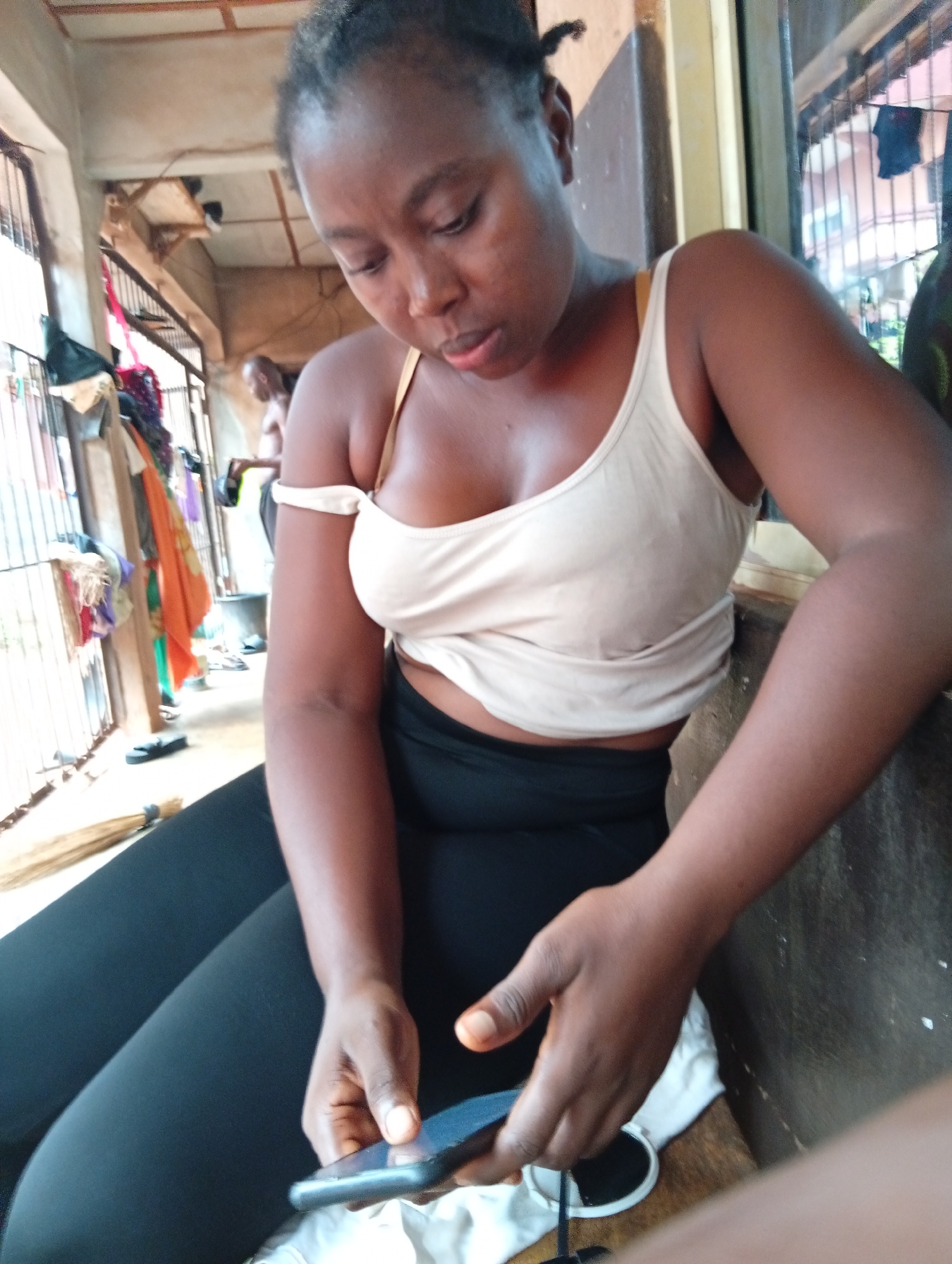 A woman is sitting down and looking at her cell phone. She is wearing a white tank top and black leggings. In the background, there is an open doorway with a person standing in the doorway. The location is Unknown, Unknown.