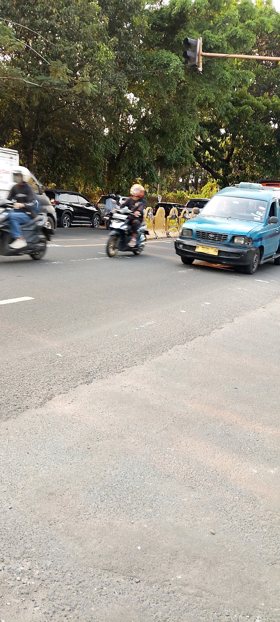 In Depok, Indonesia, at an intersection with a traffic light visible in the upper right, a blue van is turning right. Two motorcycles are also present on the road, one on the left ridden by a person in a dark jacket and blue jeans, and another on the right ridden by a person wearing a helmet with orange accents. A black SUV is parked behind the motorcycle on the left, and other vehicles are partially visible in the background. The road surface is asphalt with visible lane markings. Trees line the background of the scene.