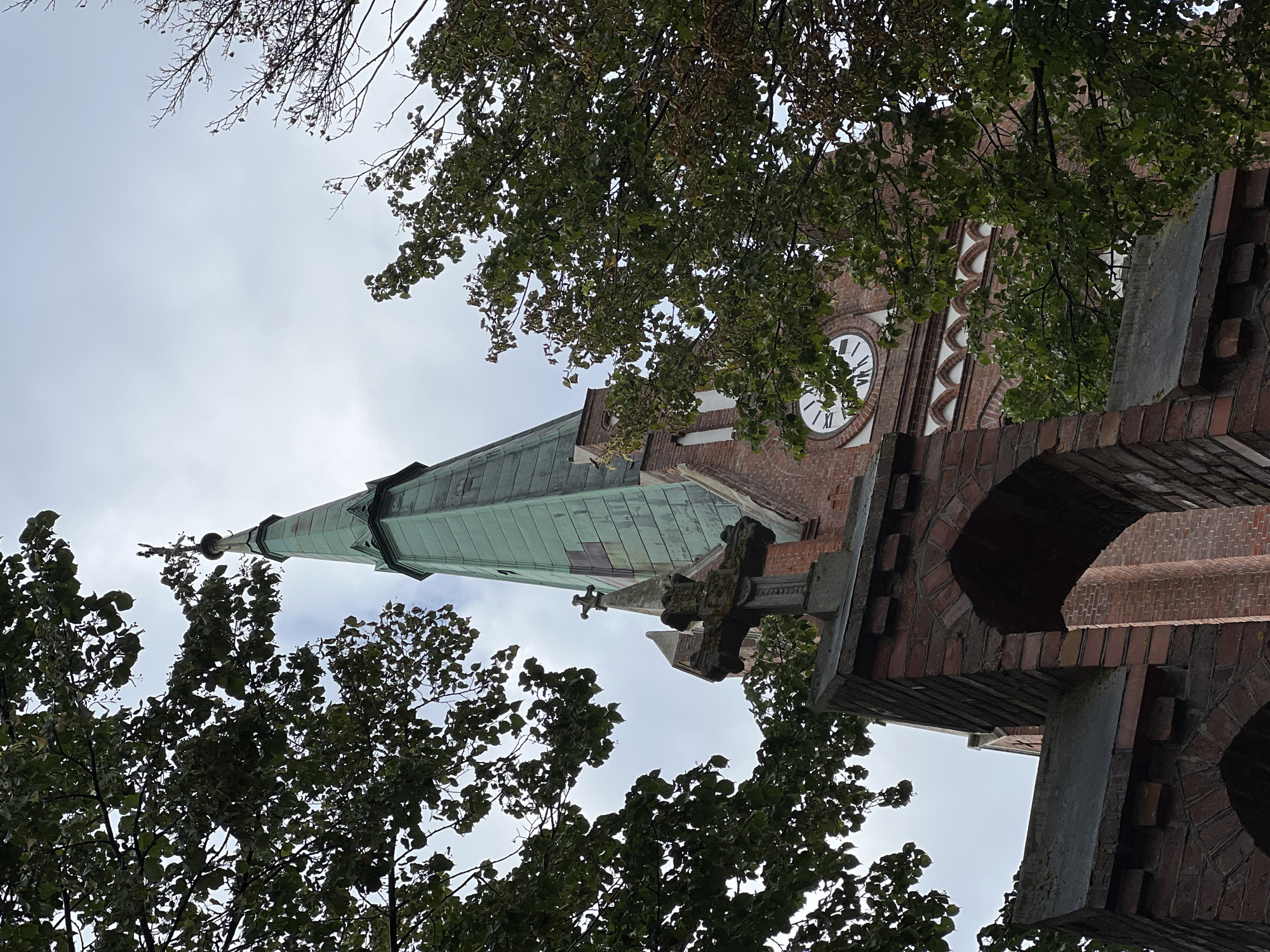 A low-angle shot captures the steeple of a red brick church in Palanga, Lithuania, partially obscured by dark green trees. The steeple, topped with a cross, is covered in green, weathered material. Below the steeple, a stone cross sits atop a brick structure with arched openings. A clock face with Roman numerals is visible on the church's brick facade. The sky is overcast.