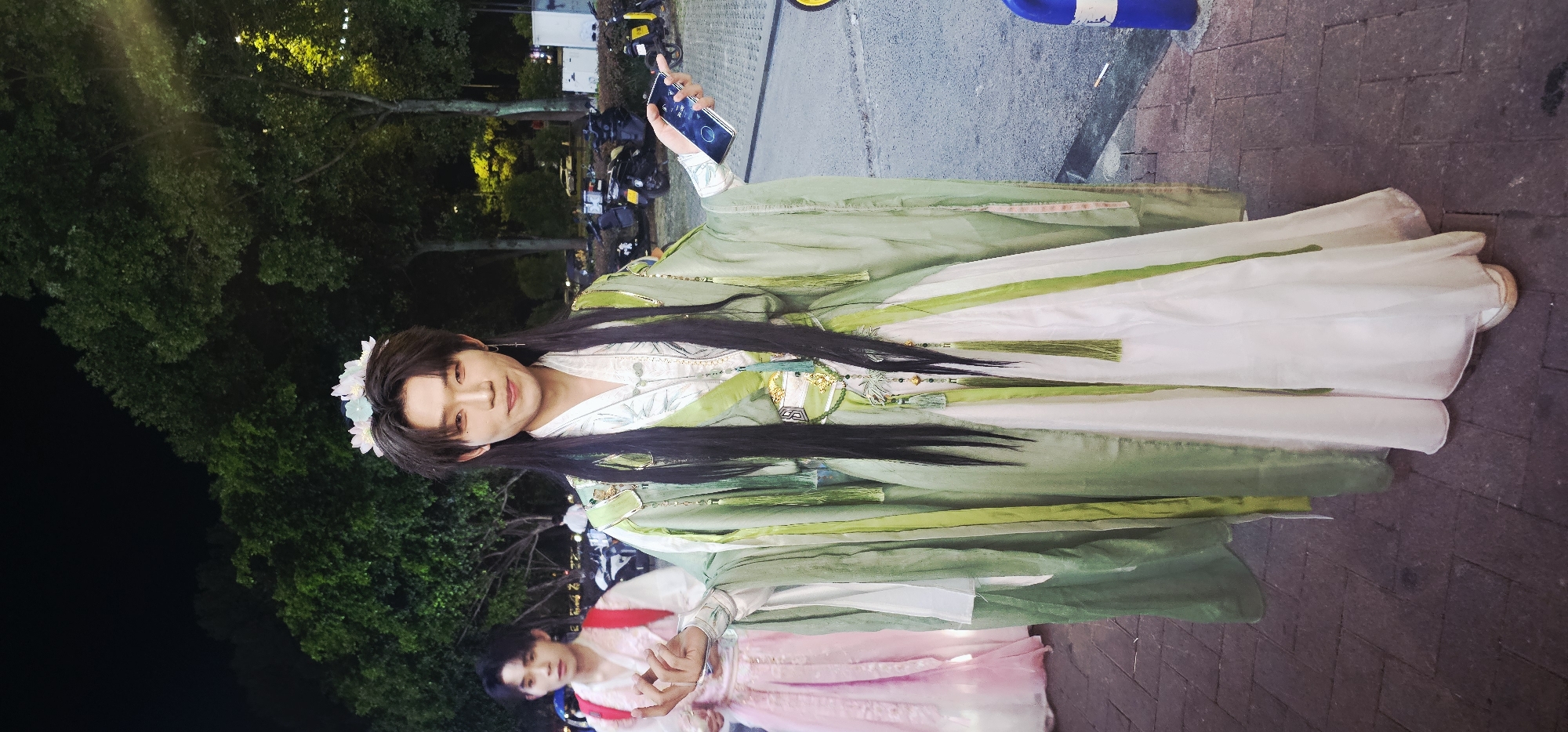 In Chongqing High-tech Zone, China, at night, a person dressed in traditional Chinese attire, featuring a light green robe with white accents and floral headwear, stands centrally, holding a smartphone in their right hand. The person has long, dark hair and is looking towards the camera with a slight smile. To the left, another individual in a pink traditional Chinese outfit is visible, facing partially away from the camera. The background shows trees with streetlights illuminating them, and several parked motorcycles are also present, indicating an urban outdoor setting. The ground is paved with brick-like tiles.