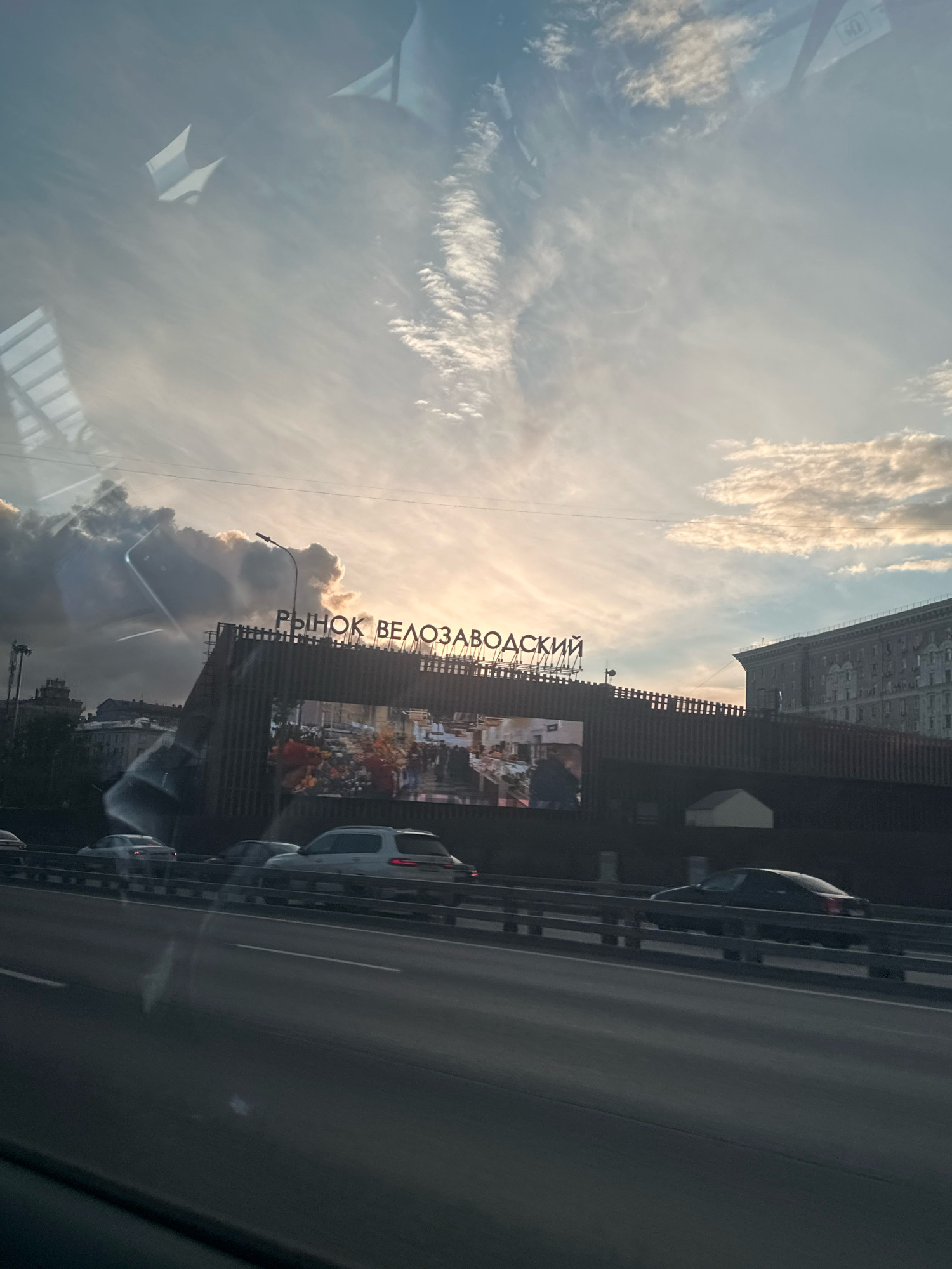 In an urban environment, likely during daylight hours as indicated by the sky and lighting conditions, a billboard displaying a lively market scene is visible. The text on the billboard, in Cyrillic script, reads "РЫНОК ВЕЛОЗАВОДСКИЙ," which translates to "Velozavodsky Market." The billboard is mounted on a structure resembling a building facade, with a dark, textured exterior. Above the billboard, the sky is a dramatic canvas of clouds, with streaks of sunlight breaking through, suggesting either sunrise or sunset. In the foreground, a multi-lane road is visible with several cars traveling in the same direction. The vehicles appear to be modern passenger cars, suggesting a contemporary setting. The overall scene captures a slice of urban life, with the prominent billboard serving as a focal point against the backdrop of the sky and traffic.