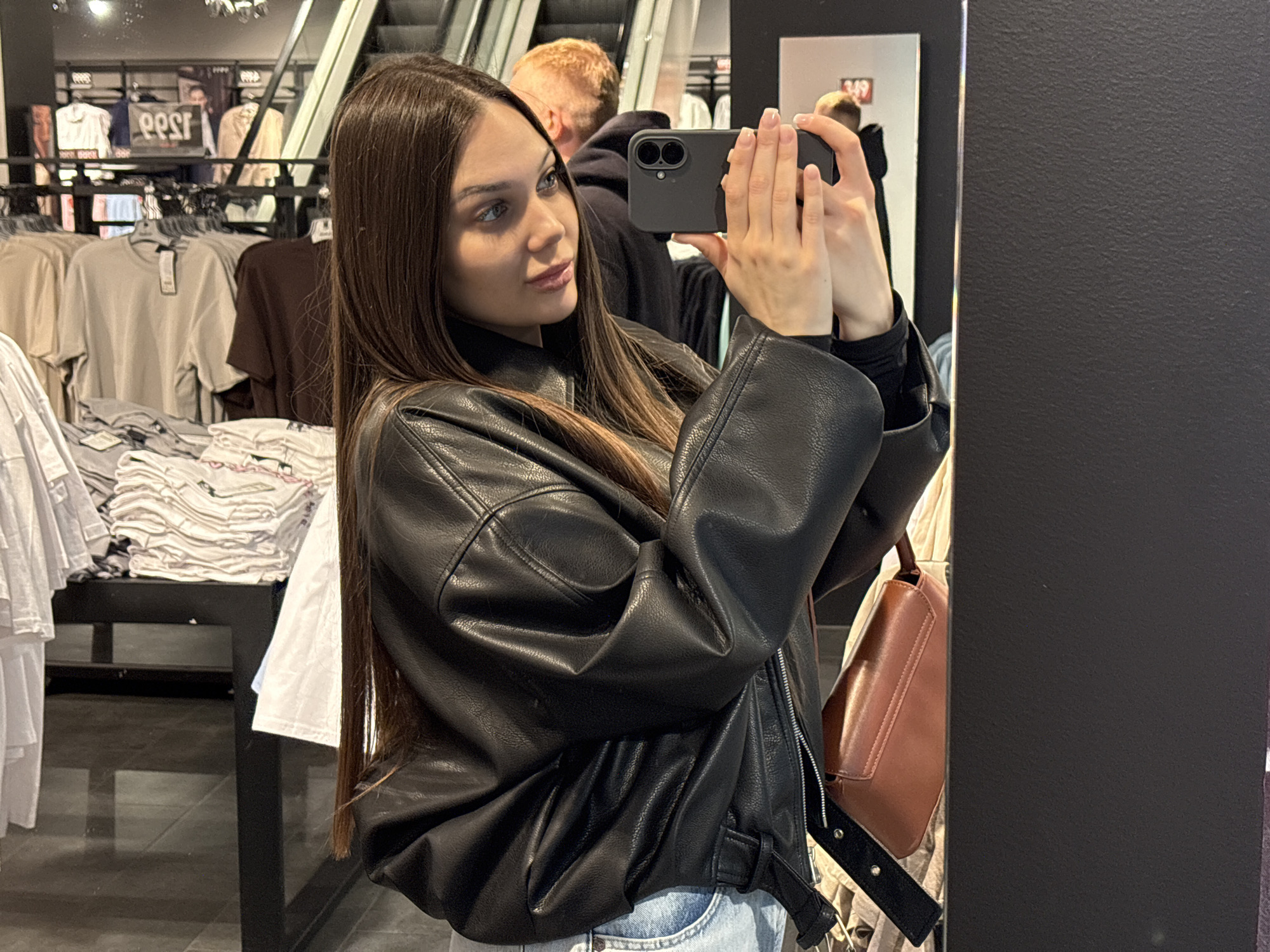 A woman with long brown hair is taking a selfie with a smartphone in front of a mirror. She is wearing a black leather jacket and blue jeans. Behind her, there are clothing racks with folded shirts and clothes hanging on hangers, suggesting a retail environment. An escalator is visible in the background. A man with red hair is partially visible in the background, slightly behind the woman. A brown handbag is hanging nearby. The scene appears to be in a clothing store, potentially located in the city of Yekaterinburg, Russia.