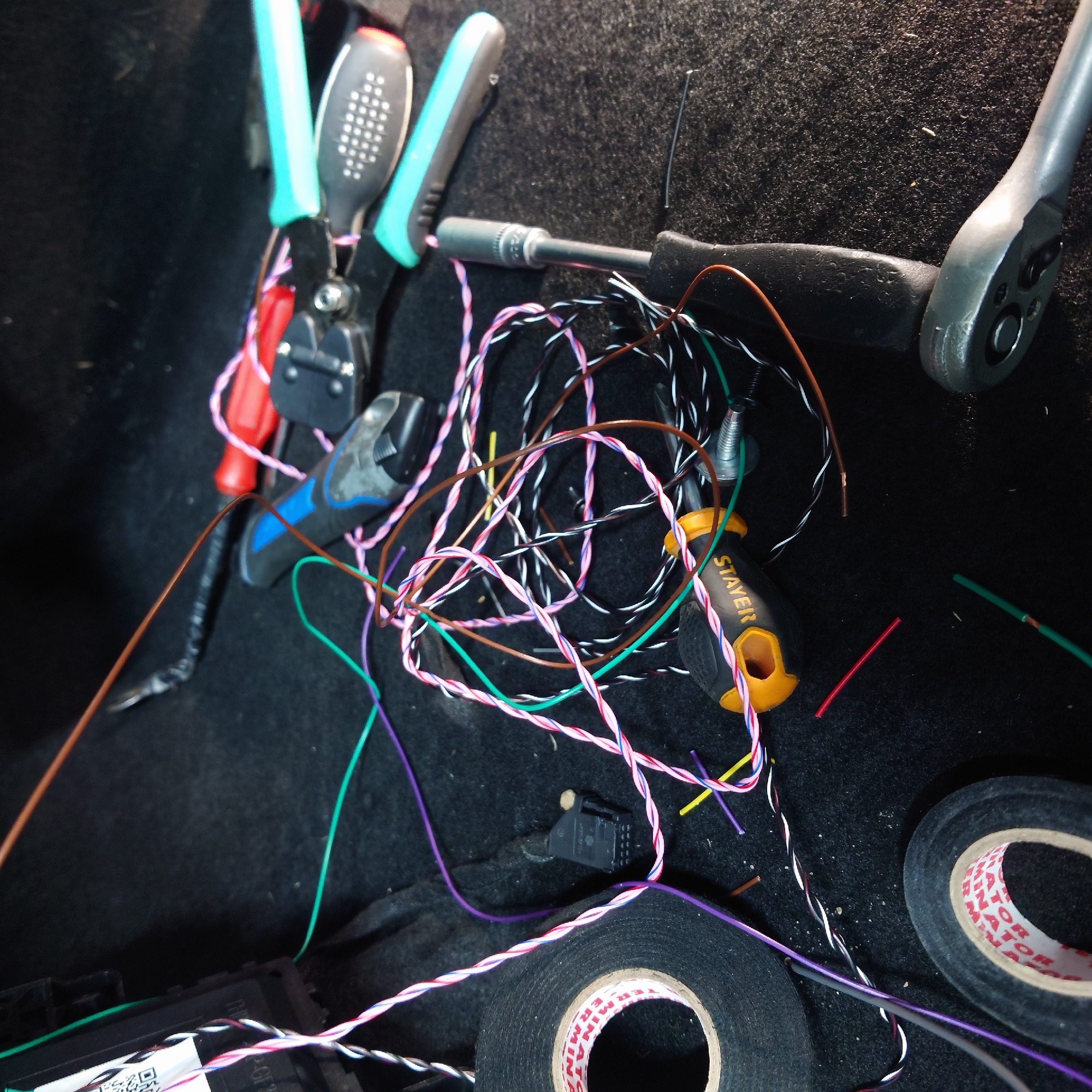 The image shows a close-up view of car wiring work, likely in the trunk or under a seat, given the dark, carpeted surface. Various tools and electrical components are scattered on the surface.
Dominant objects include a tangle of multi-colored wires (white, pink, green, brown, and purple), electrical tape rolls, a set of wire strippers with teal and black handles, a pair of pliers with blue and black handles, a socket wrench with a silver head and black handle, and a yellow-handled screwdriver with the brand name "STAYER" visible on its side. There is also a black electrical connector with multiple pins.
The wires appear to be either in the process of being connected, disconnected, or organized. The presence of tools like strippers and pliers suggests active work is being done on the wiring. The electrical tape suggests that connections are being insulated.
There are no people visible in the image. The lighting appears to be artificial, as there are no strong shadows or indications of natural light sources. The setting is indoors, within a vehicle.
No specific time of day or weather conditions can be inferred. The overall impression is one of a DIY automotive repair or installation project.
Visible text includes the brand name "STAYER" on the screwdriver handle and lettering on the tape rolls which appears to be "MINATOR" or similar. There is also a QR code and some text on a label attached to a black electronic component at the bottom left.