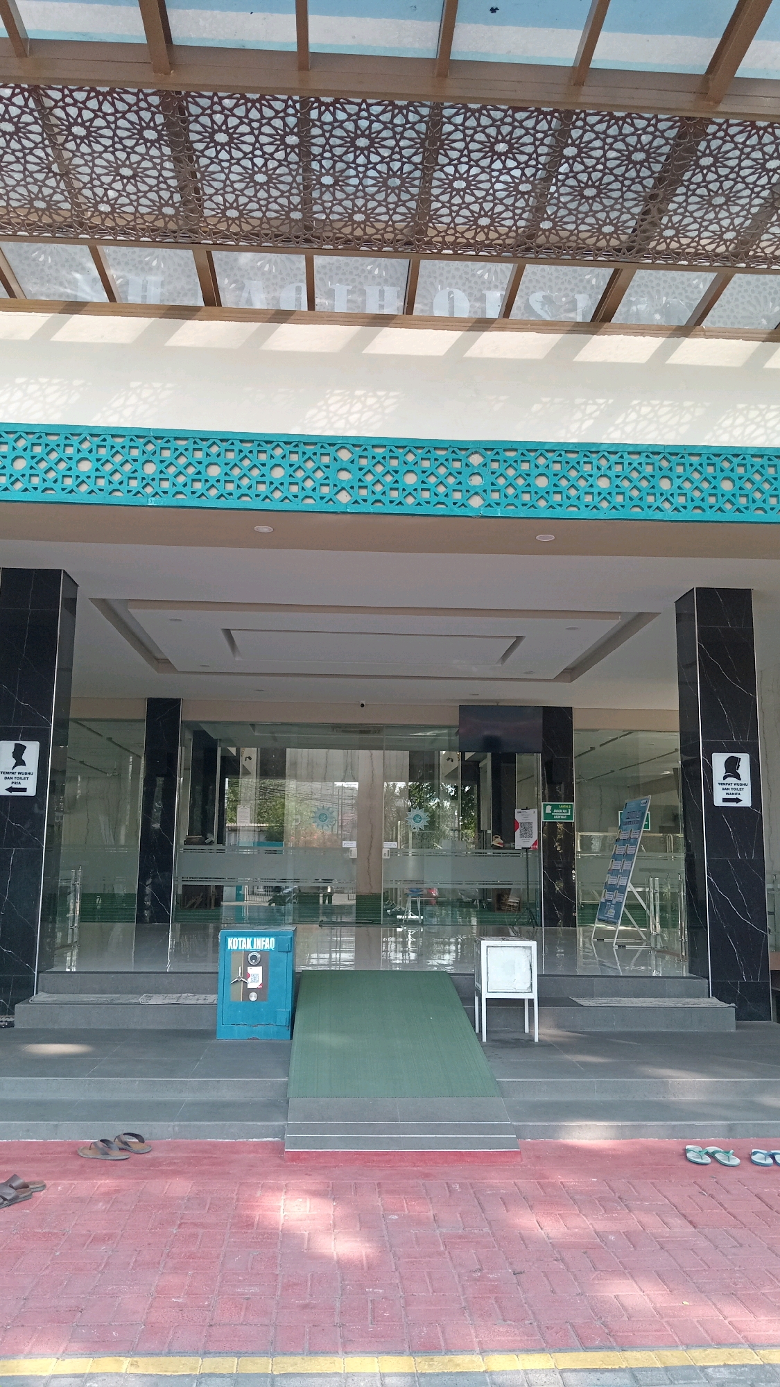 The image shows the entrance to a building, possibly a mosque, with distinctive Islamic geometric patterns on the ceiling and a turquoise decorative border. Two black marble-clad pillars flank the glass entrance. On the left pillar, a sign with a silhouette of a person and text indicates a designated area. To the right of the entrance, a similar pillar has a sign with a silhouette of a woman and text, likely indicating a women's area. In front of the entrance, there is a ramp leading up to the building. To the left of the ramp, a teal-colored donation box labeled "KOTAK INFO" is visible. A white table-like structure stands to the right of the ramp. The ground in front of the building is paved with red bricks. The overall environment suggests an urban or semi-urban setting in Randuagung, Indonesia, likely a public or religious building with accessibility features.