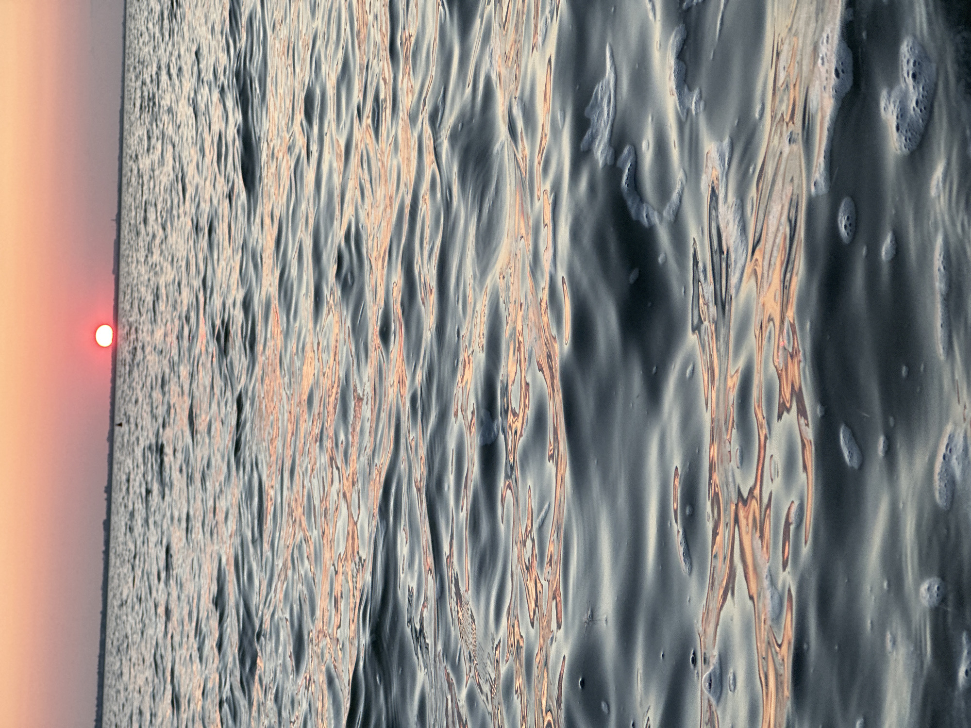 A calm body of water is shown with gentle waves, reflecting the soft hues of a sunset. The sun, a brilliant orb of orange and red, is low on the horizon, casting a warm glow across the sky and the water's surface. A distant shoreline with a few indistinct structures and a small buoy are visible. The water itself is dark gray, with subtle ripples creating patterns of light and shadow. Patches of white foam are scattered across the foreground, indicating movement or perhaps the presence of small waves. The overall impression is one of tranquility and natural beauty. This scene is not specific to Kokshetau, Kazakhstan, as the image depicts a generic sunset over water.