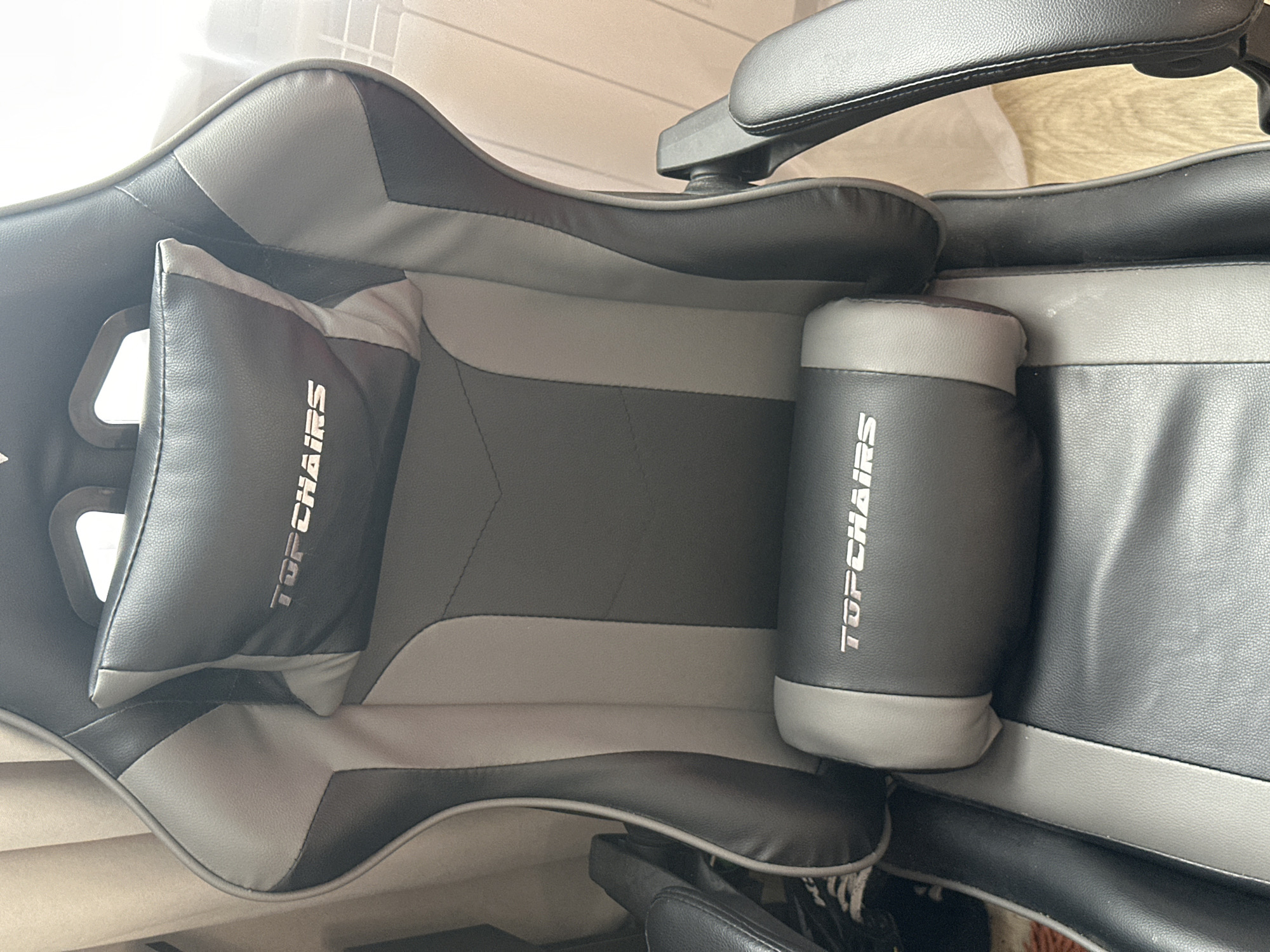 The image shows a close-up, head-on view of a black and gray gaming chair. The chair has a high back with adjustable headrest and lumbar support pillows, both of which are emblazoned with the silver logo "TOPCHAIRS". The chair is made of faux leather and features contrasting gray and black panels. The lighting suggests it is daytime, with natural light coming from the right side of the frame, casting faint shadows. The background is out of focus but appears to be an indoor setting with curtains on the left and what looks like a radiator and wooden flooring on the right. No people are visible in the image. The location in Birobidzhan, Russia cannot be determined from the visual information provided.