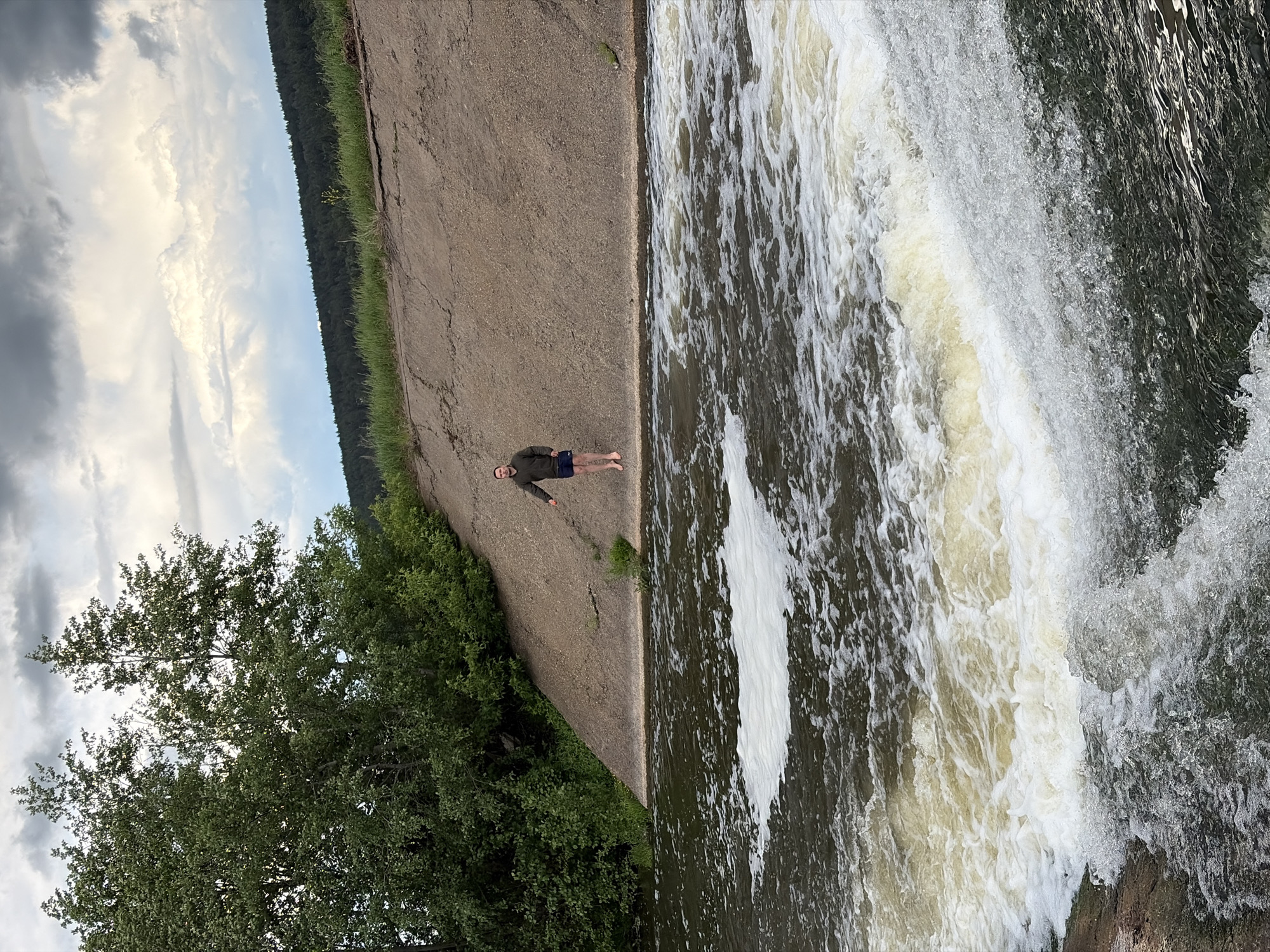 The photograph depicts a waterfall and adjacent concrete structure in Nizhnii Vishur, Russia. A person stands barefoot on the concrete near the waterfall's edge. The setting includes a mix of natural and man-made elements, with trees and sky visible in the background.