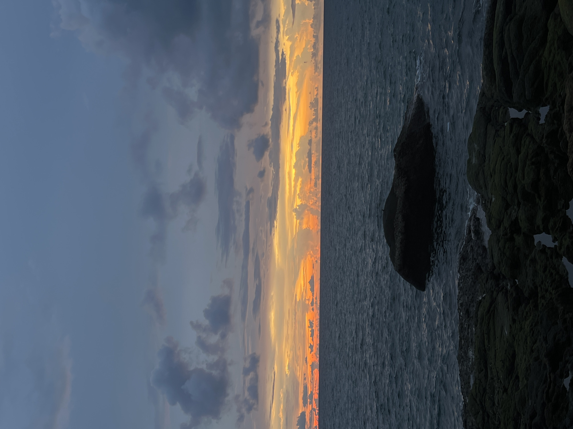 The image depicts a rocky coastline at dusk, with the sun setting over the ocean. The sky is filled with dramatic clouds, ranging from dark gray to vibrant orange and yellow hues. The sea is a deep blue, with gentle waves lapping against the shore. In the foreground, dark, moss-covered rocks are visible, with some tide pools reflecting the twilight sky. A large, dark boulder sits in the water, partially submerged. The overall mood is serene and peaceful. This scene is likely located on the coast of Karon, Thailand, known for its beautiful beaches and stunning sunsets.
