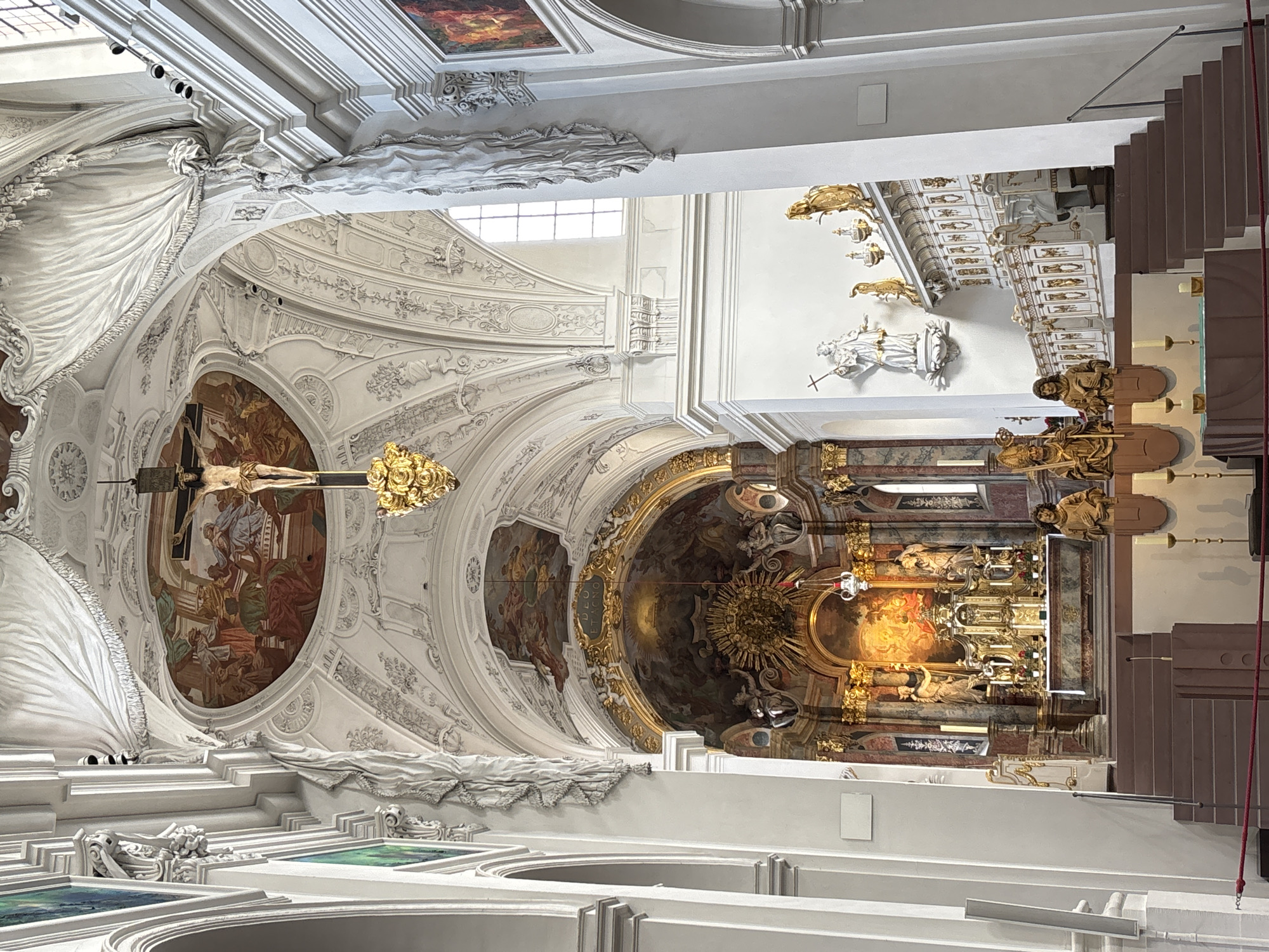 The image shows the interior of the Würzburg Residence, specifically the Kaisersaal (Imperial Hall). The hall is elaborately decorated with Baroque architecture and artwork. In the foreground, a grand staircase leads up to the altar area, which is adorned with intricate gold-leaf ornamentation and paintings. Above the altar, a fresco adorns the ceiling, depicting a historical scene. Suspended from the ceiling by a golden chain is a crucifix, a central element of the religious setting. Sculptures of figures, likely saints, are placed along the walls and on pedestals, adding to the grandeur of the space. Sunlight streams in from tall windows, illuminating the ornate details of the hall.