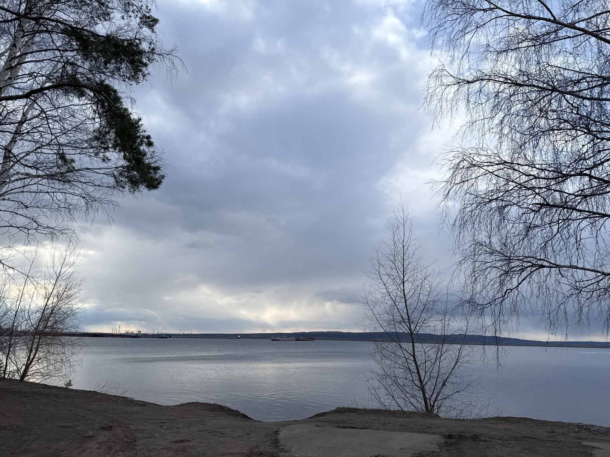The image captures a tranquil, expansive waterscape under a dramatic, cloudy sky. In the foreground, a dark, earthy bank forms the bottom edge of the frame, hinting at a shoreline or path. Bare trees frame the scene on both sides. To the left, a prominent birch tree with its characteristic white bark and intricate dark, leafless branches stands tall, intermingled with the dark green foliage of a coniferous tree. On the right, delicate, bare branches of another tree extend into the frame, creating a natural archway.
The central part of the image is dominated by a vast body of calm, grey water, which subtly reflects the light from the sky. In the middle distance, a long, low-profile barge or boat is visible on the water, moving slowly across the scene. The distant horizon reveals a faint landmass, possibly a far shore, with subtle hints of industrial structures like cranes or power lines silhouetted against the lighter part of the sky.
The sky is heavily overcast with various shades of grey clouds, suggesting an impending storm or a generally gloomy, diffused light. The overall mood is quiet and contemplative, perhaps evoking a sense of late winter or early spring due to the bare trees. There are no people visible, and no discernible text in the image.