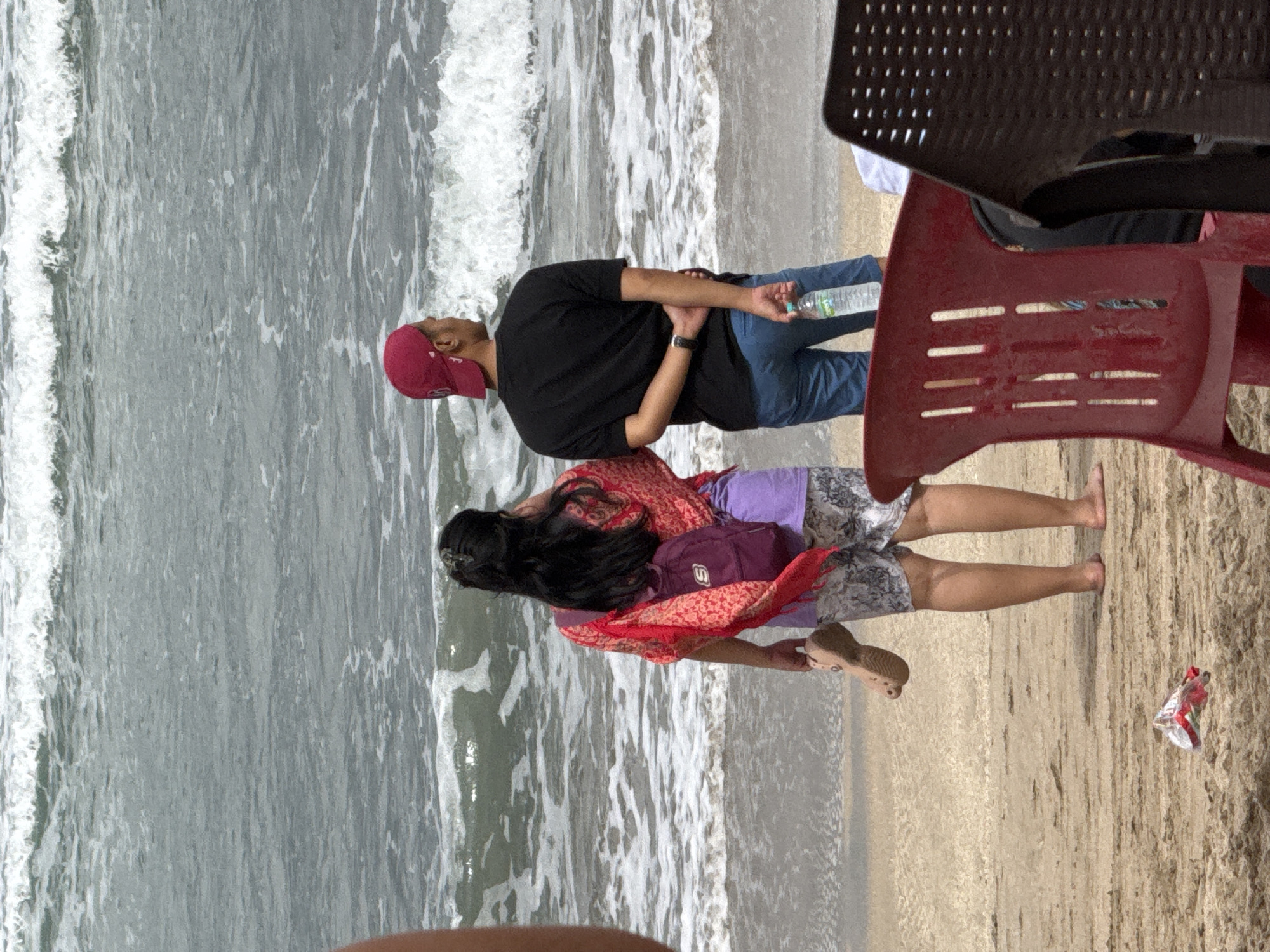 In Kuta, Indonesia, two individuals stand on a sandy beach facing the ocean. A person with dark hair, wearing a red patterned shawl over a purple garment and patterned shorts, holds a sandal in their right hand. They also have a purple backpack on their back. Beside them, a person wearing a red baseball cap, a black t-shirt, and blue shorts holds a bottle of water in their right hand. Their arms are clasped behind their back. In the foreground, red and dark brown chairs are partially visible. The ocean, with visible waves, stretches out behind the people, indicating a coastal setting.