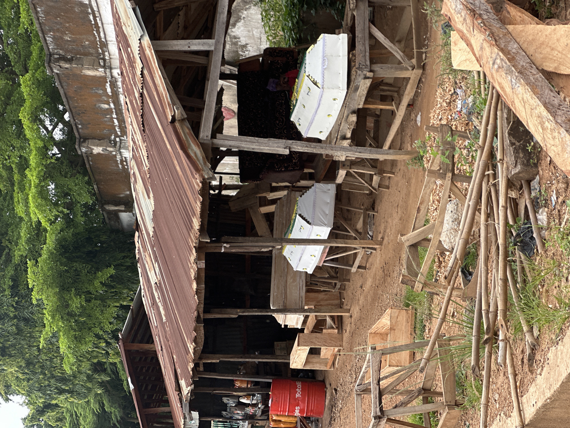 The image depicts an outdoor workshop, likely a coffin maker's establishment, situated in a somewhat cluttered and rustic environment. The workshop appears to be constructed from rough-hewn wood and corrugated metal roofing, showing signs of age and weathering. Green foliage is visible in the background, suggesting an outdoor setting perhaps in a rural or semi-rural area.
The primary objects in the scene are coffins. Two are particularly prominent, decorated with white fabric and possibly some yellow accents. One coffin is open, revealing what appears to be a person dressed in dark fabric, possibly a shroud or funeral attire, lying inside. Another coffin is visible below it, also covered in white fabric. Various pieces of wood, some processed and some in log form, are scattered around the workshop, indicating ongoing construction or material storage. A red fuel barrel, labeled "TotalE", is also present, along with other miscellaneous items and debris.
The overall impression is one of a functional, working space. The lighting suggests it is daytime, possibly midday given the ambient light. The setting appears to be in Akwanga, Nigeria, as indicated by the provided location context. There are no explicit cues about the weather, but the presence of green vegetation implies it is not a dry season. The scene is devoid of any overt emotional expression, focusing on the practicalities of the workshop and its contents. The visible text "TotalE" on the red barrel is the only legible text in the image.