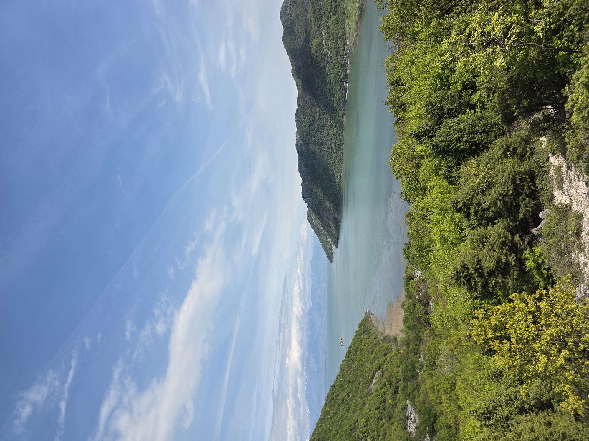 The image shows a view of Skadar Lake in Godinje, Montenegro. The lake is surrounded by green mountains and dense vegetation. The sky is light blue with streaks of clouds. In the distance, mountains are visible through a haze.