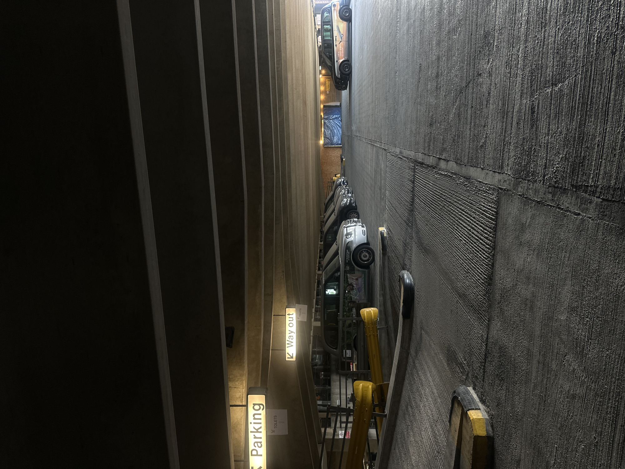 A high-angle view, slightly rotated counter-clockwise, captures the interior of a parking garage in London, United Kingdom. The left side reveals vertically stacked, dark gray, concrete slabs that form a wall with a series of rectangular indents. Light fixtures with signs reading "Parking" and "Way Out," along with directional arrows, are visible between the slabs. The right side features a rough, textured concrete wall, with several layers of different finishes. A yellow and gray guardrail, along with a few tires acting as bumpers, are attached to the wall. Several parked cars are visible between the two walls, ranging from silver sedans to black cabs. A blue tarp is visible in the distance. The artificial lighting casts shadows and illuminates the scene.