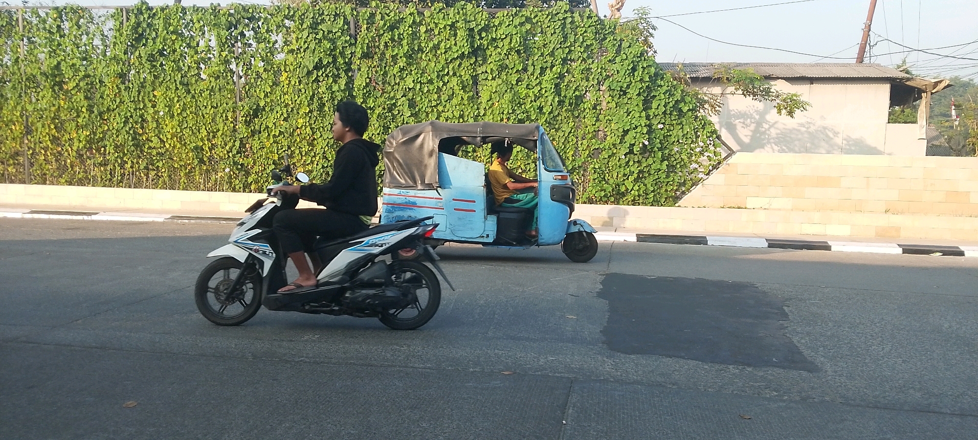 A motorized tricycle and a motorcycle are traveling on a road in Satriamekar, Indonesia. The tricycle is light blue with a black canopy and has two passengers. The driver, wearing a yellow shirt and green pants, is at the controls. A passenger in a light brown shirt is seated behind the driver. The motorcycle is white and black and is being ridden by a young man wearing a black hooded jacket and black pants. He is wearing flip-flops. Both vehicles are moving in the same direction. The road has a dark asphalt surface with some lighter patches. To the left of the road is a low, light-colored retaining wall, and behind that is a dense green hedge or wall of vegetation. Further to the right, a light brown tiled wall is visible, with a simple structure with a corrugated metal roof behind it. The sky is bright and appears to be late afternoon or early morning, with soft lighting and shadows.