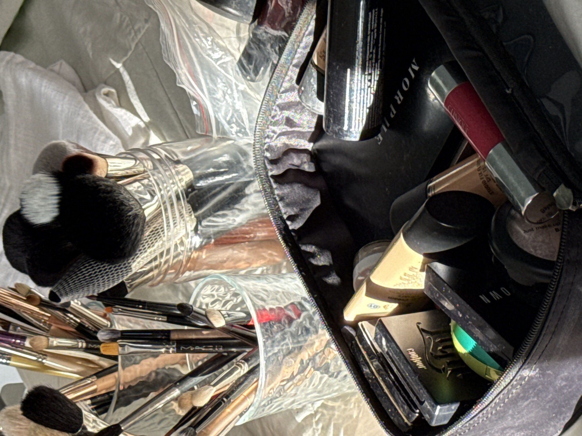 The image shows a cluttered scene with makeup items. Two clear glass jars are filled with makeup brushes, with the brush heads visible at the top of the jars. To the right, a black makeup bag is partially open, revealing various makeup products including a "Morphe" item, a Clinique lipstick, foundation, and palettes. The items are packed tightly within the bag. The background consists of a white crumpled cloth or fabric, and a clear plastic bag. The location and city are unknown.
