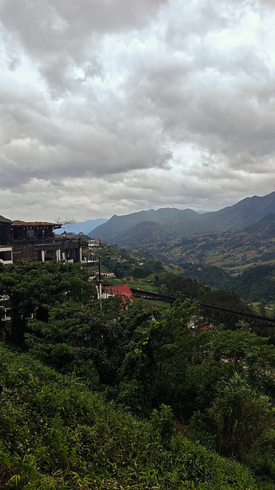 The scene depicts a mountainous landscape under a cloudy sky. The foreground is filled with lush green foliage and vegetation, partially obscuring a red-roofed structure and a dark-colored walkway or bridge.
In the midground, there is a multi-tiered building with a wooden exterior and glass windows, built into the hillside. Further back, a valley stretches out, dotted with small villages and terraced fields, leading to a series of rolling mountains in the distance. The sky is overcast with dense, grey clouds, suggesting a humid or rainy climate.
The location is likely Sa Pa Ward, Vietnam, based on the terraced landscape and architectural style. There are no people visible in the scene.