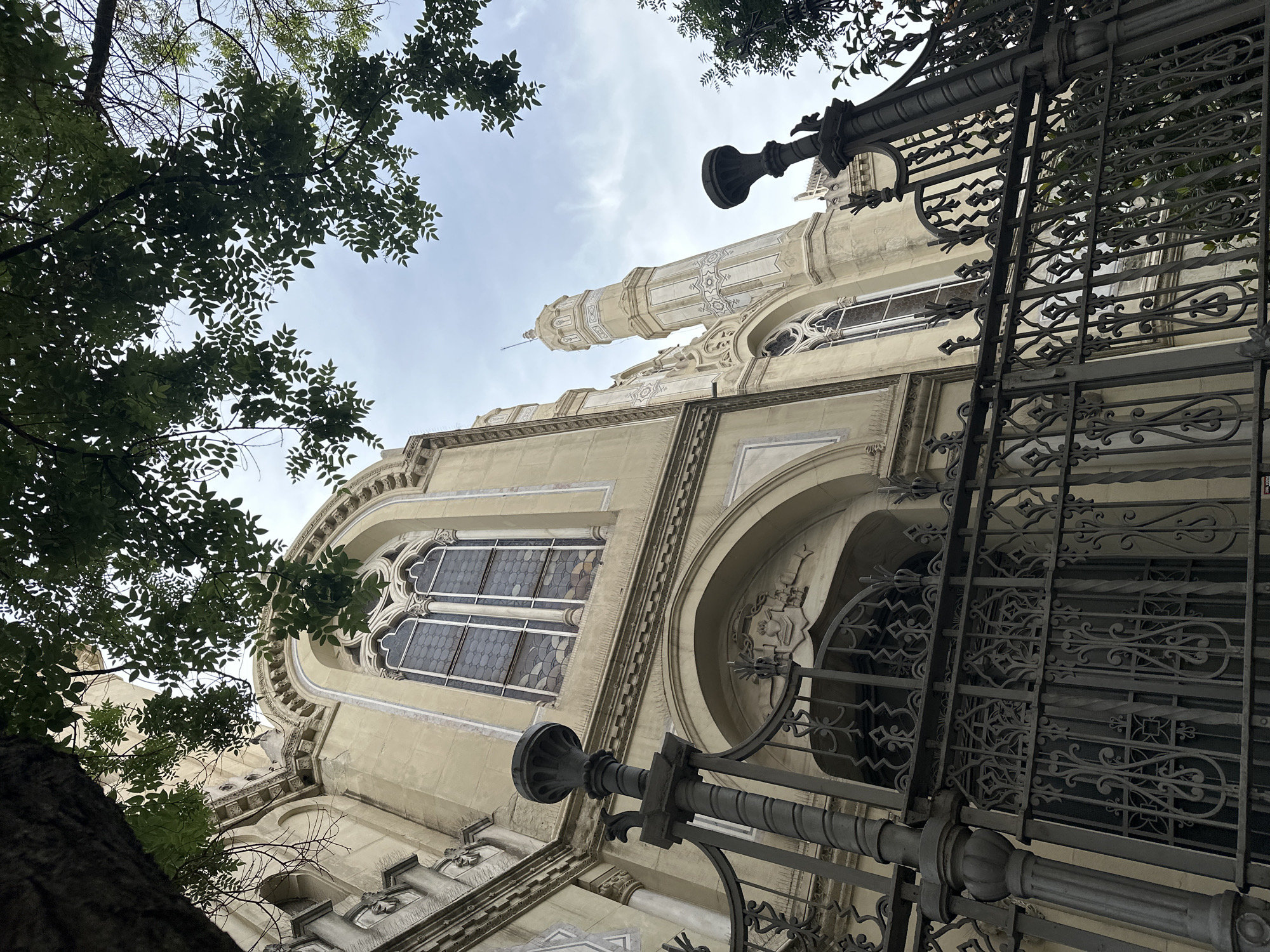 The image shows a low-angle view of the Mezquita Central de Madrid (Madrid Central Mosque) in Madrid, Spain. The mosque's architecture includes a minaret, arches, and patterned windows. The building is framed by tree branches and a section of ornate iron fencing. The sky is overcast.