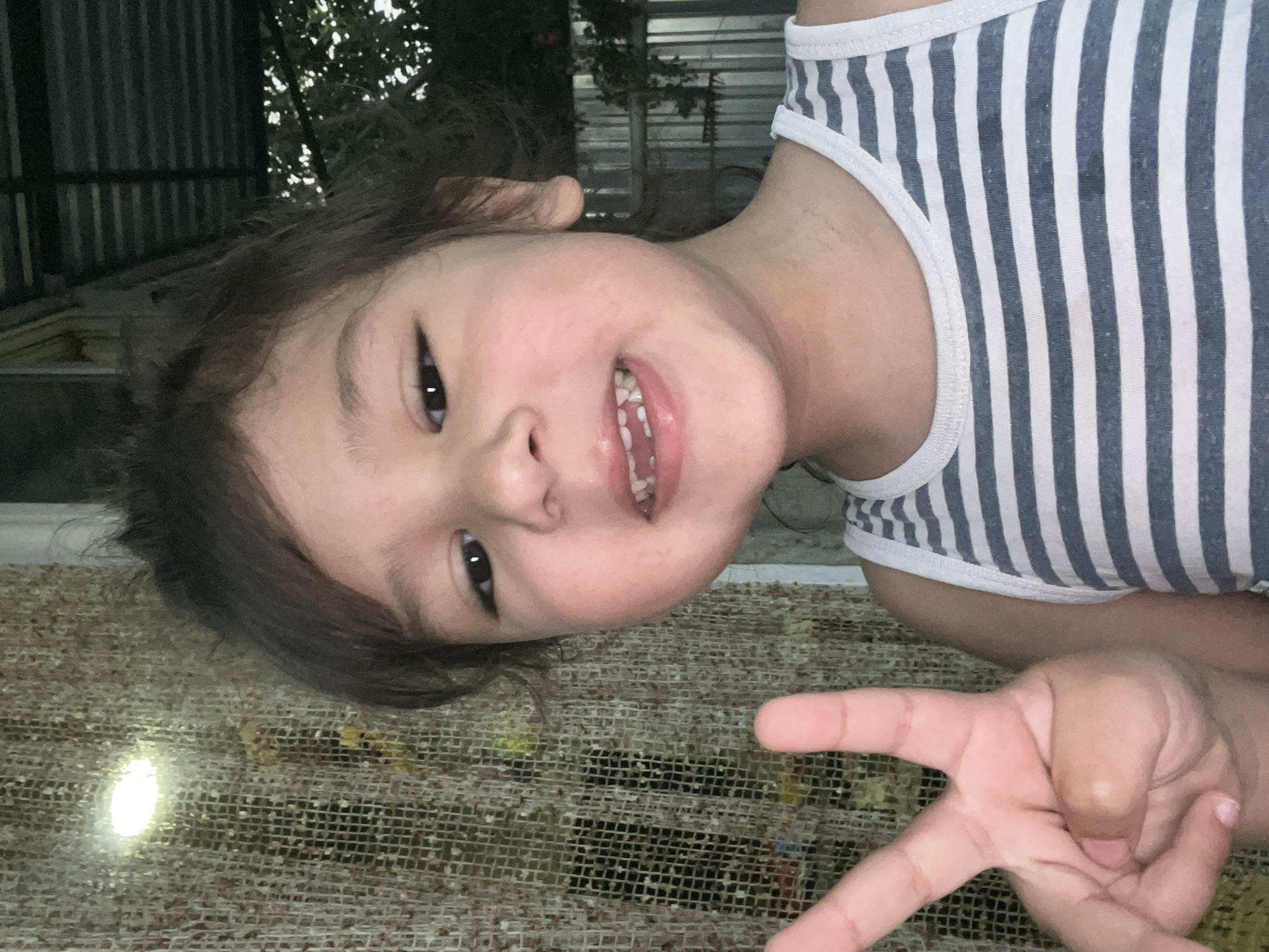 An outdoor, close-up, eye-level shot captures a young child making a peace sign with their left hand. The child, facing the camera, has dark hair and is wearing a gray and white striped tank top. Their mouth is open, revealing several teeth. To the left of the child, a textured, beige-colored surface is visible, with a bright light source illuminating a portion of it. In the background, there is a metal fence and foliage, suggesting an outdoor setting. The scene does not provide any specific details or visual cues to determine its location in the city of Tbilisi, Georgia.