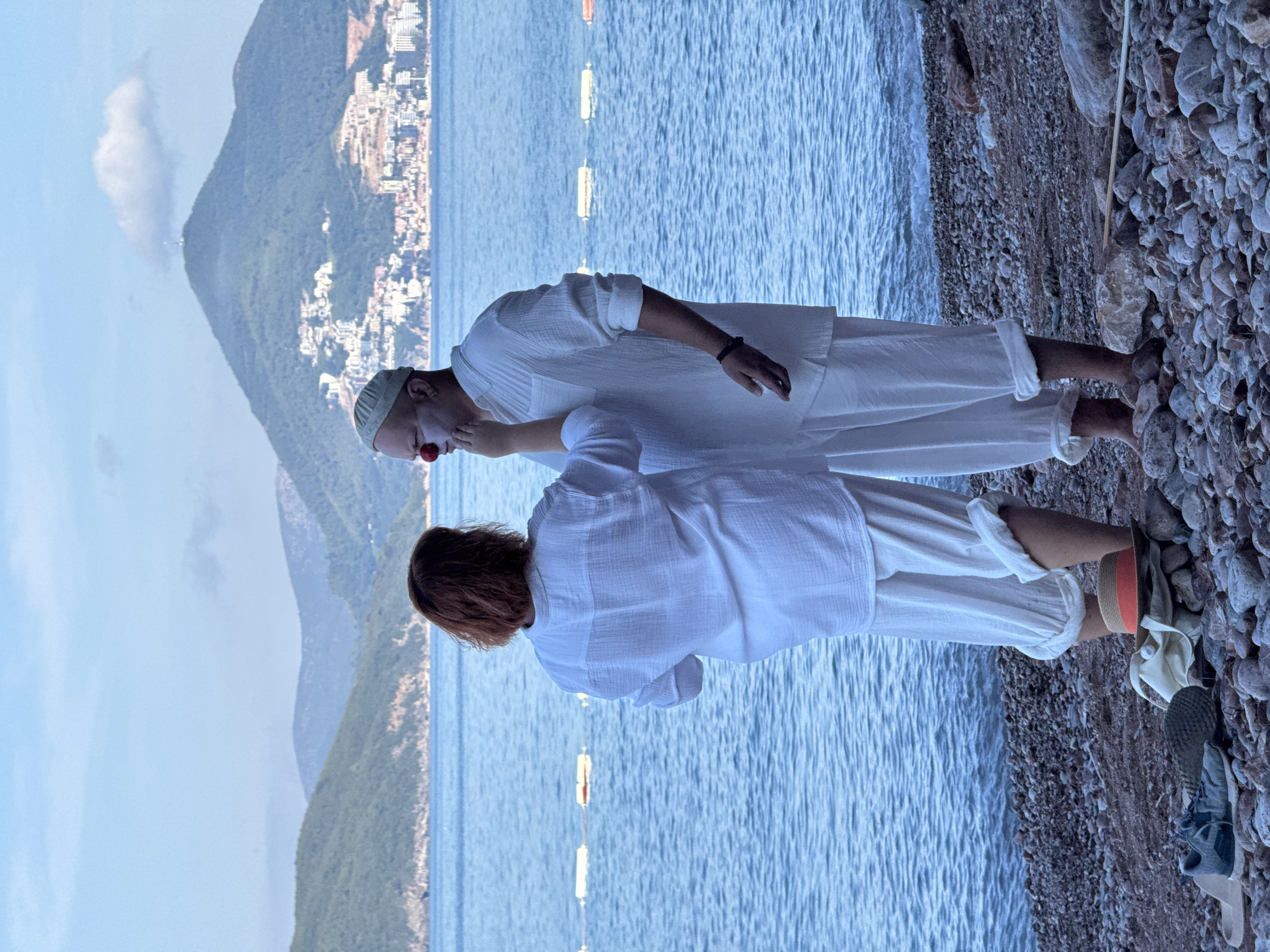 A woman with auburn hair, facing away from the camera, applies makeup to a man's face. The man is wearing a white cap and has a red clown nose and white face paint. Both are dressed in loose-fitting white clothing. They stand on a pebble beach with the blue sea behind them. In the distance, green, tree-covered hills rise, with a city built on the slopes and a tower on the summit of the tallest peak. The city appears to be Sveti Stefan, Montenegro, as the distinctive island town is visible in the background. The sea has buoys floating on its surface. On the beach, near the man's feet, are a straw hat with an orange band, a canvas bag, and a pair of athletic shoes.