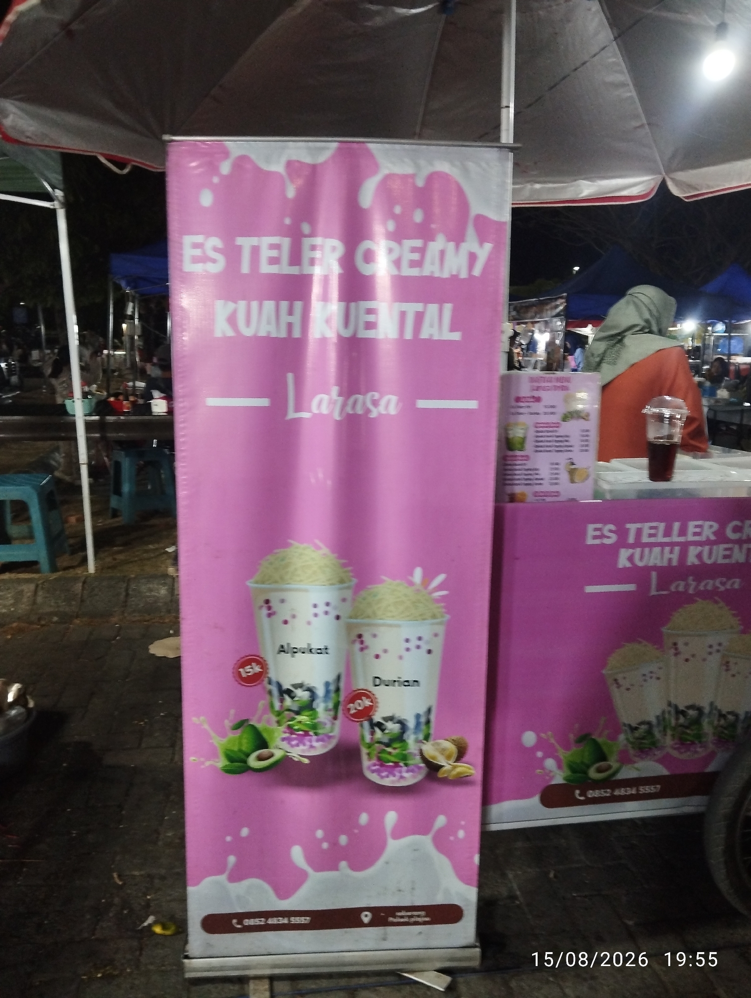 The image shows a street food stall at night in a city, likely in Southeast Asia, given the text on the banners. The stall features a large pink banner with white text advertising "ES TELUR CREAMY KUAH KENTAL" and "Larasa", which translates to "Creamy Egg Es with Thick Broth" and "Larasa" (likely a brand name). Below this text are illustrations of drinks in cups, with labels "Alpukat" (avocado) and "Durian". There are prices indicated as "15k" and "20k" (likely 15,000 and 20,000 Indonesian Rupiah). Another smaller pink banner to the right also displays similar advertising. In the background, there are tents and people, suggesting a market or fair. There is an umbrella overhead, and some stools are visible. A person wearing a hijab and an orange top is visible behind the counter. The ground is paved. A timestamp "15/08/2026 19:55" is present in the lower right corner.