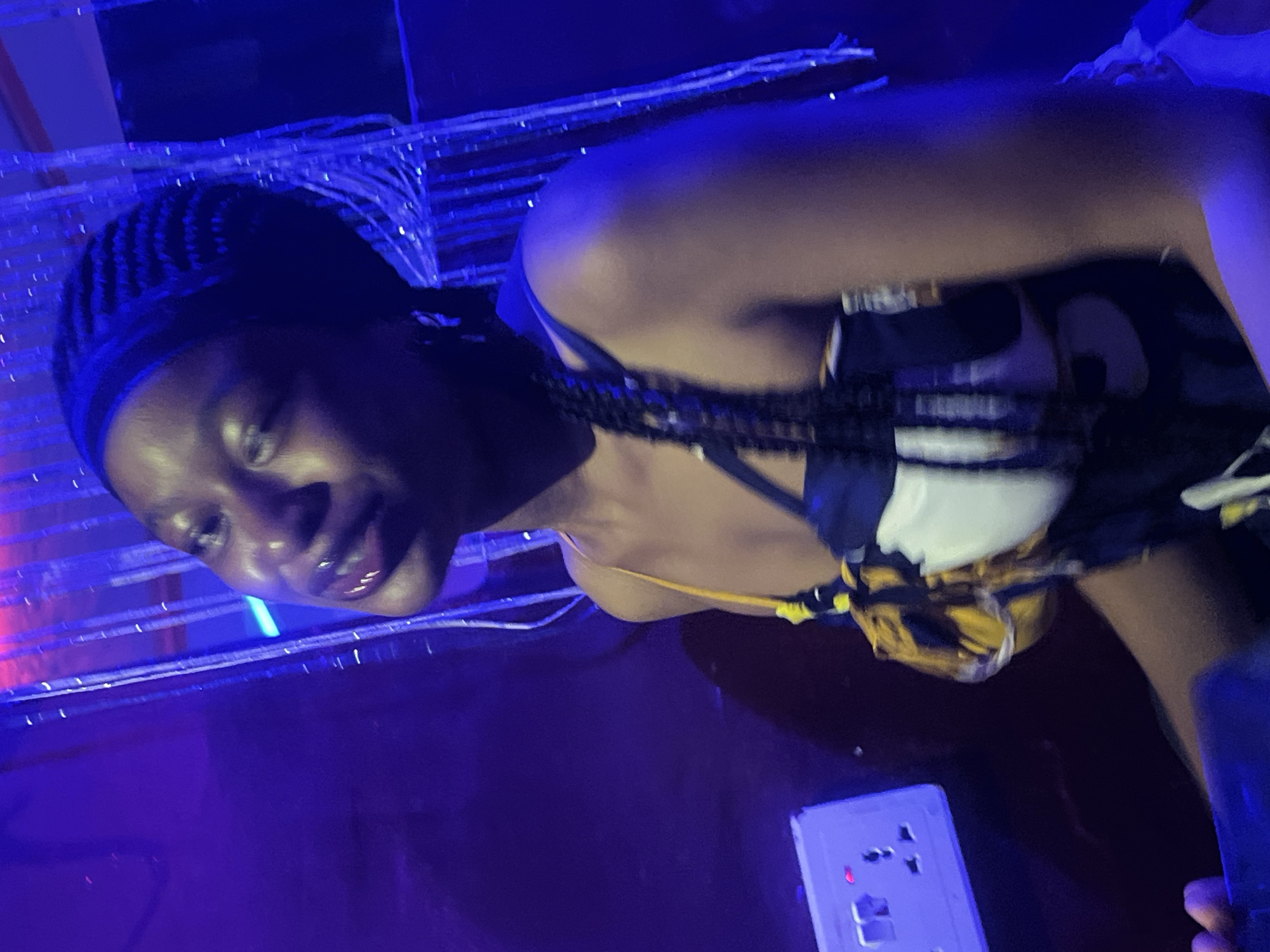 A young Black woman with braided hair and wearing a patterned dress is pictured from the chest up, angled towards the viewer. She is lit by purple and blue lights, with her face partially in shadow. To her left, a white electrical outlet is visible against a dark background. Behind her, shimmering vertical strands hang, illuminated by the colored lighting. The setting appears to be an indoor space, potentially a club or lounge, with a distinct lack of urban elements typical of Durumi, Nigeria, as the background is dominated by decorative lighting and drapes, not cityscapes or recognizable Nigerian landmarks.