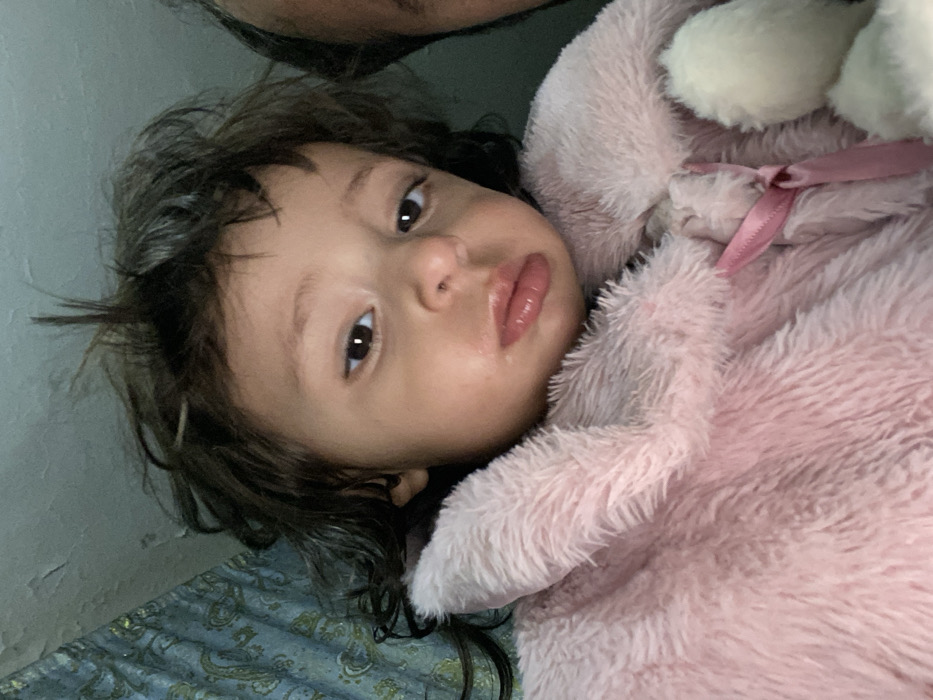 A young child with brown hair and eyes rests on a paisley patterned surface. They are snuggled in a light pink, fluffy blanket or stuffed animal with a satin ribbon. The child's face is slightly damp, with full lips and a focused gaze. Part of an adult's face is visible in the upper left corner. The background includes a wall with visible imperfections.