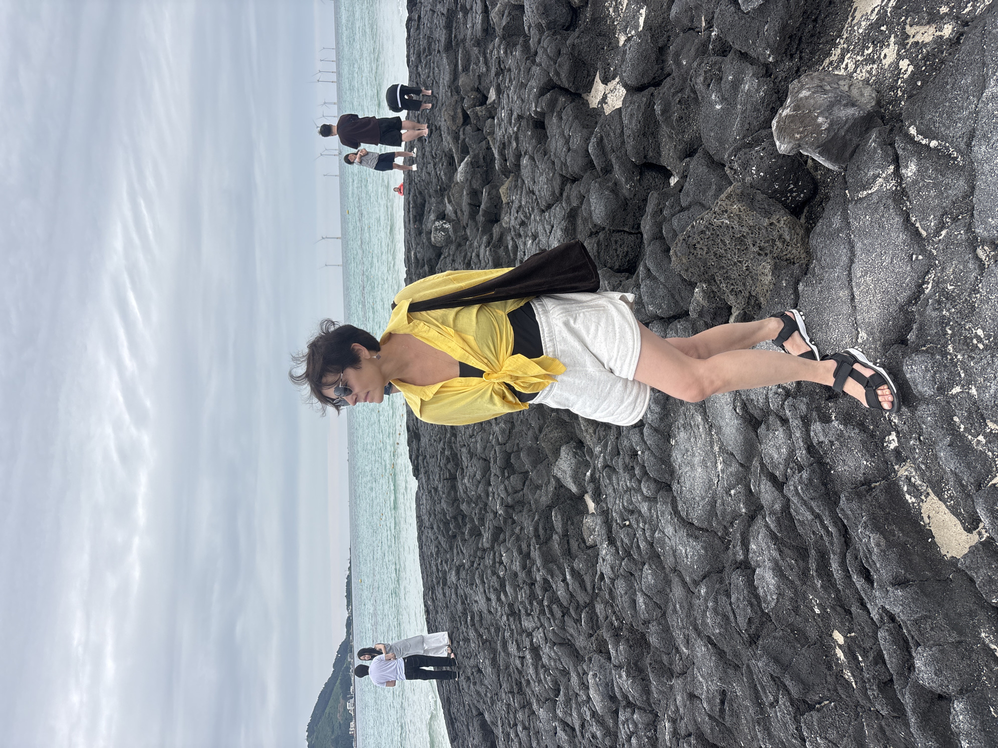 The image shows a woman in a yellow shirt and white shorts standing on a rocky shoreline. She is wearing sunglasses and black sandals. Behind her, the ocean is visible with several wind turbines in the distance. The sky is overcast, suggesting a cloudy or possibly rainy day. The rocks appear to be dark volcanic rock, common to coastal areas of Jeju, South Korea. The overall scene depicts a casual outing to the coast, with the woman posing for a photo while enjoying the view. In the background, other people are visible near the water's edge, and a small town or settlement can be seen on a distant hillside. The mood of the image is relaxed and serene.