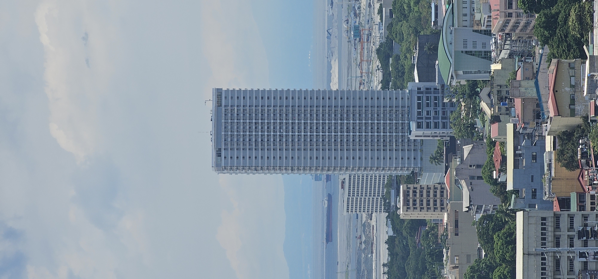 The image shows a cityscape with a tall, rectangular skyscraper as the dominant feature, centrally located. The skyscraper is light-colored with a grid-like pattern of windows and balconies. In the background, a body of water with a large cargo ship is visible, along with distant hazy mountains. The foreground and mid-ground reveal a dense urban environment with various buildings of different heights and architectural styles, interspersed with lush green trees. A construction site with cranes and piles of materials is also present in the mid-ground. The sky is partly cloudy with a bright, overcast atmosphere. The scene appears to be a daytime view of an urban area, possibly a port city, with the specific mention of Makati, Philippines, suggesting it is a view within that city's metropolitan area.