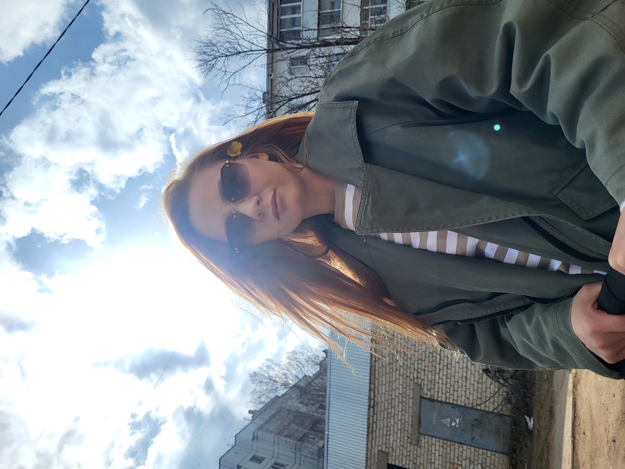 A woman is outdoors, seemingly in the city of Chaikovsky, Russia, based on architectural styles. She wears sunglasses, a green jacket, and has a yellow flower tucked behind her ear. Her hair is reddish-brown. She is possibly taking a selfie. The background includes a cloudy sky, a brick building with a metal door, and a taller building with bare trees nearby. The lighting is bright, suggesting a sunny day. The image is rotated slightly.