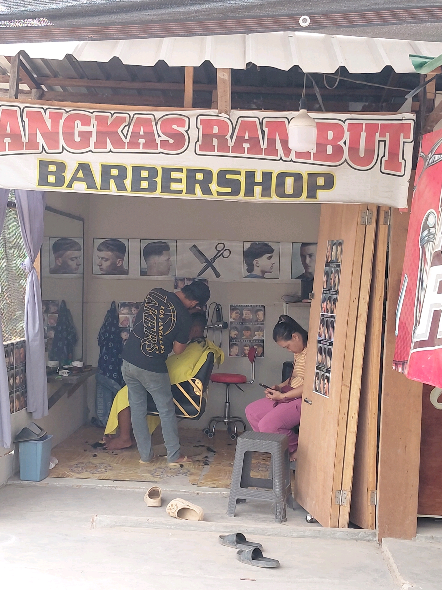 In the city of Persiapan Mulya Raya, Indonesia, a barbershop is depicted, its name, "ANGKAS RAMBUT BARBERSHOP," prominently displayed on a banner. The shop appears to be an open-air establishment, with a simple roof structure and wooden doors.
Inside, a barber, wearing a black t-shirt and blue jeans, is in the process of cutting a client's hair. The client is seated in a barber chair, draped with a yellow cloth. The barber is focused on his work, holding clippers and tending to the client's head.
On the wall behind the barber, a series of framed pictures display various hairstyles, likely for customer reference. To the left of the barber, a woman is seated, engrossed in her mobile phone. She wears a striped shirt and pink pants.
The floor of the barbershop is covered with scattered hair clippings, indicating recent activity. To the left of the shop, a blue trash bin is visible. Outside the shop, on the ground, a pair of beige slippers and a pair of black flip-flops are present. The overall environment suggests a functional, no-frills barbershop serving the local community.