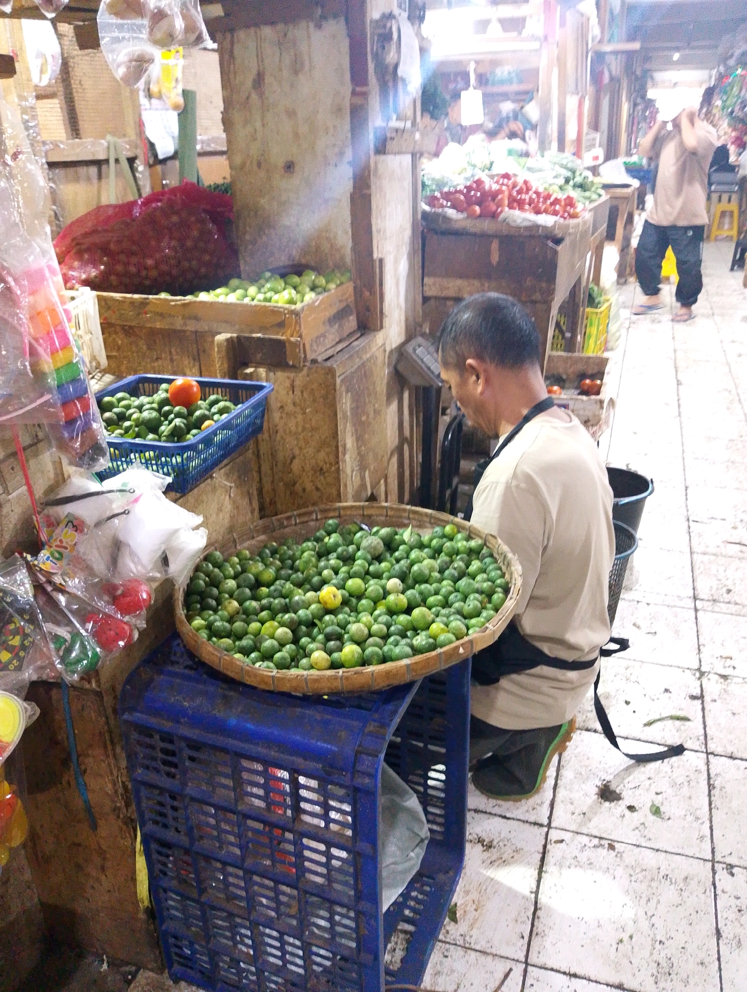 In a bustling outdoor market, likely in Lembang, Indonesia, a man kneels beside a blue plastic crate. He is wearing a light-colored t-shirt, a black apron, and dark green rubber boots. His attention is focused on a large, round woven basket overflowing with small, green citrus fruits, possibly calamansi. The man appears to be sorting or preparing the fruit.
The crate beneath the basket holds more produce, indistinct in the shadows. To the left of the crate, various items are displayed, including colorful plastic toys, bags of produce, and a red net bag filled with what seems to be red onions. In the background, stalls are laden with fresh produce such as tomatoes and leafy greens. Another person, partially visible, stands in the background near a stall. The market floor is tiled, and scattered debris suggests a busy trading environment. The lighting is bright, indicating it is daytime.