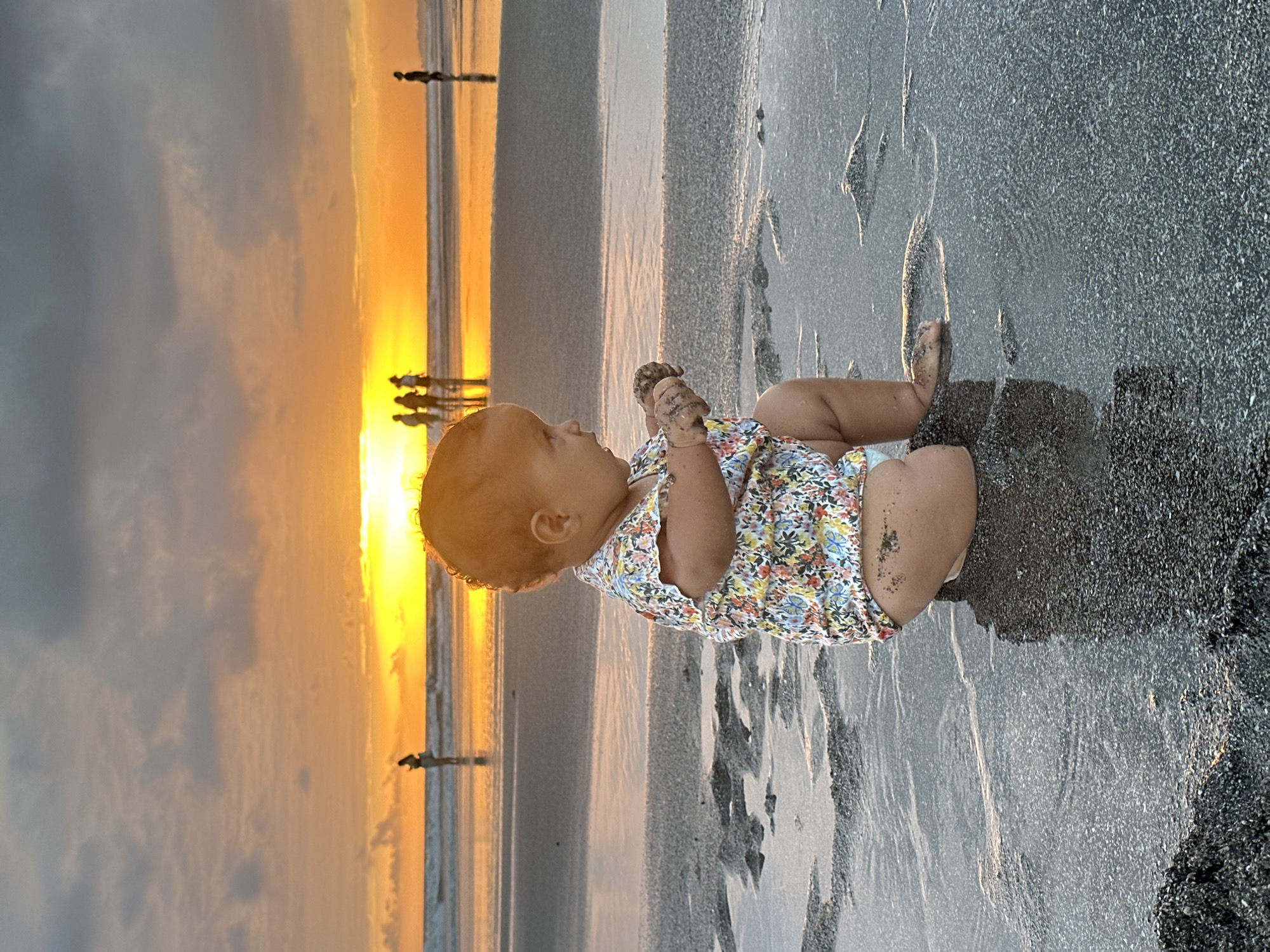 A baby is crouching on a sandy beach in North Kuta, Indonesia, at sunset. The baby is wearing a colorful floral romper and is playing with sand in their hands. In the background, several figures can be seen walking along the shoreline, silhouetted against the vibrant orange and yellow hues of the setting sun. The ocean waves gently lap at the shore, reflecting the warm light of the sky. The sand is wet and glistening in places, with small puddles of water. The overall scene is peaceful and serene, capturing a moment of childhood innocence against a beautiful natural backdrop.