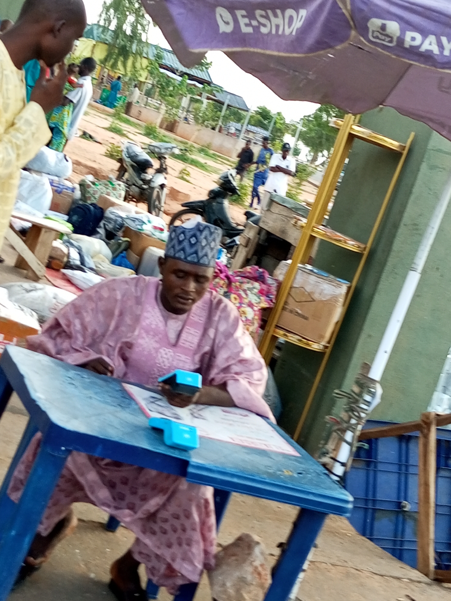 In the city of Gombe, Nigeria, a man wearing a pink kaftan and a patterned cap sits at a blue plastic table. He is looking down at a small blue device in his hand, seemingly operating it. The table has papers and other small items on its surface. The background is an outdoor market setting with a purple umbrella overhead advertising "E-SHOP" and "PAY". Several motorcycles are parked behind the table, and various goods are stacked around, suggesting a bustling commercial area. To the right of the man, a yellow shelf unit is visible, holding boxes. The ground is dirt.