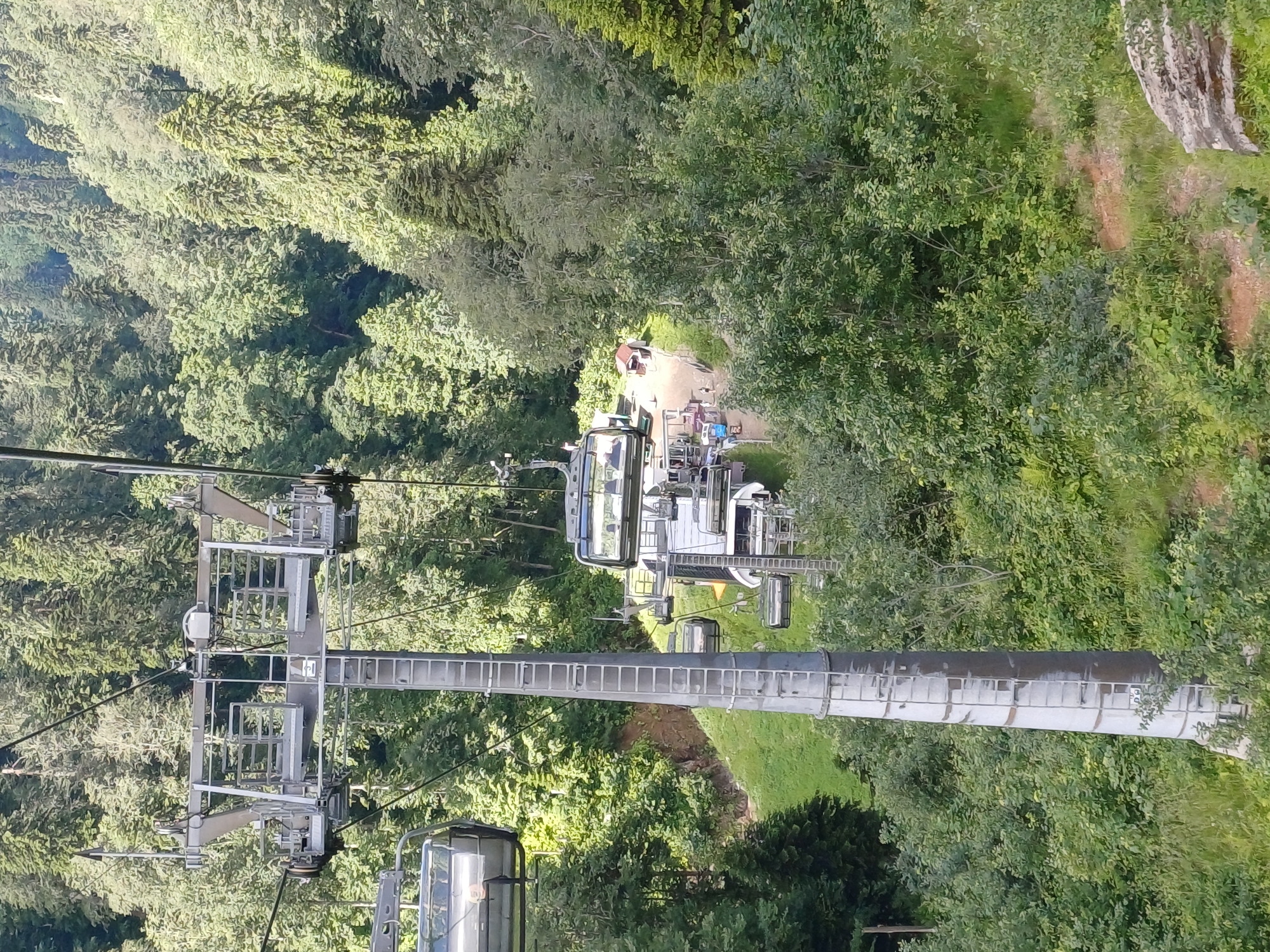 This is a photograph taken from a cable car, looking down at a cable car station and a forested mountainside in Estosadok, Russia. The cable car tower is in the foreground, with several cable cars visible on the cables. The station has multiple people and some small buildings. The surrounding environment is a dense forest with a lot of green foliage. The overall impression is one of a recreational area within a natural mountain setting.