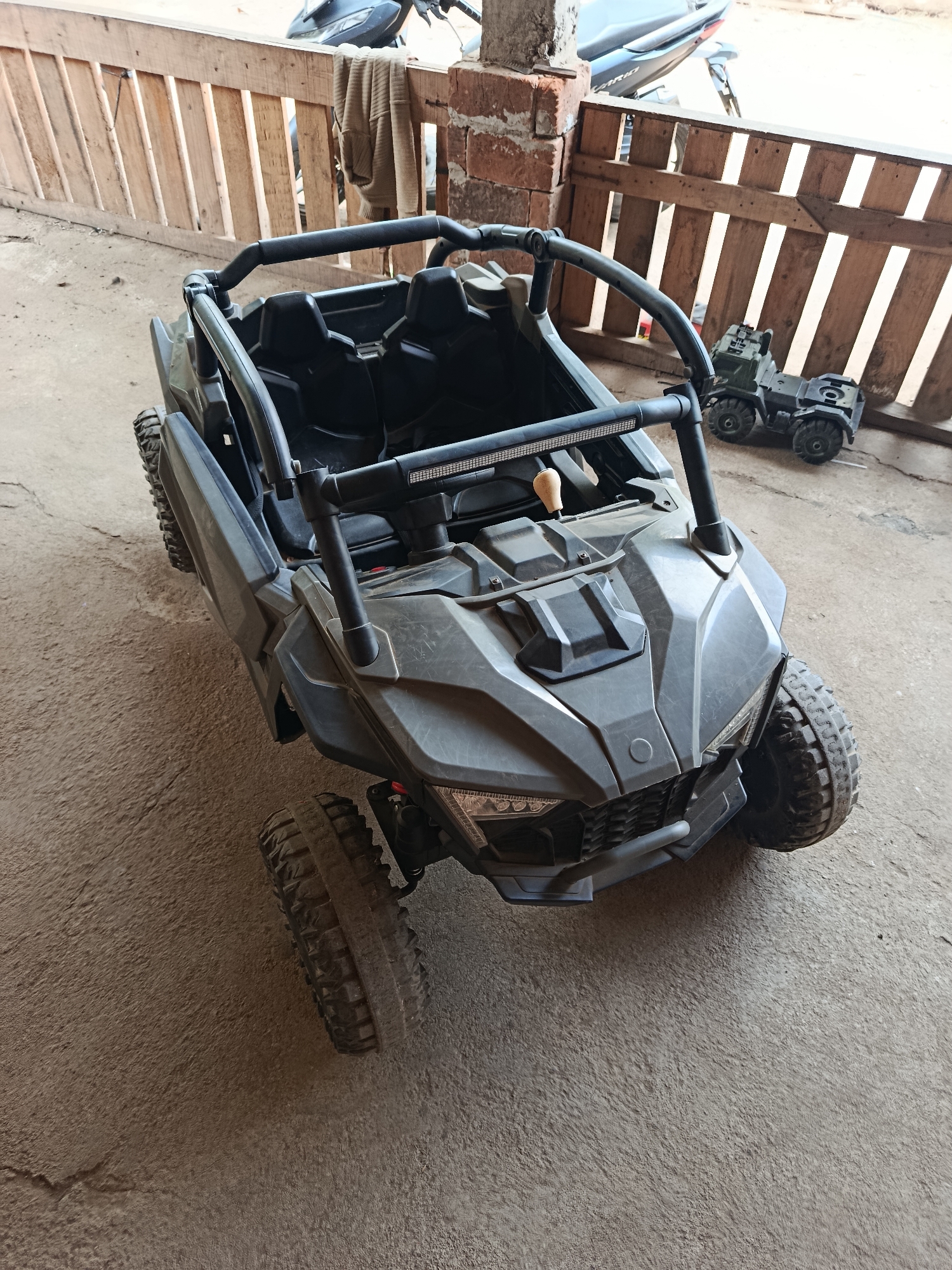 In Sedayu, Indonesia, a dark gray children's ride-on car resembling a UTV or dune buggy is parked on a concrete surface. The car has a roll cage, two black seats, and a front light bar. In the background, a wooden fence is visible, and behind it, a portion of a motorcycle and a small toy truck are also present. The scene appears to be outdoors, possibly in a yard or garage.