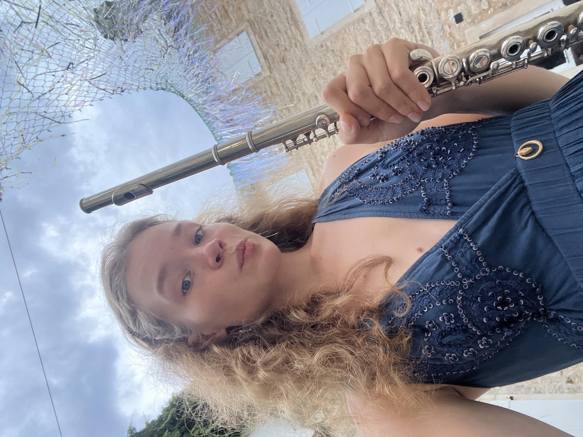 In Budva, Montenegro, a young woman with wavy blonde hair is captured in a low-angle, outdoor selfie. She is wearing a dark blue, pleated dress adorned with intricate dark blue embroidery and sequins. She holds a silver flute vertically in her right hand, with her fingers positioned on the keys. Her left hand is not fully visible. Her facial expression is a pout, with her eyes looking directly at the camera. The background features a stone building with white shutters and a cloudy sky. A decorative net with shimmering threads hangs from above, adding a festive element to the scene. The overall setting suggests a performance or an event in a historic European town.