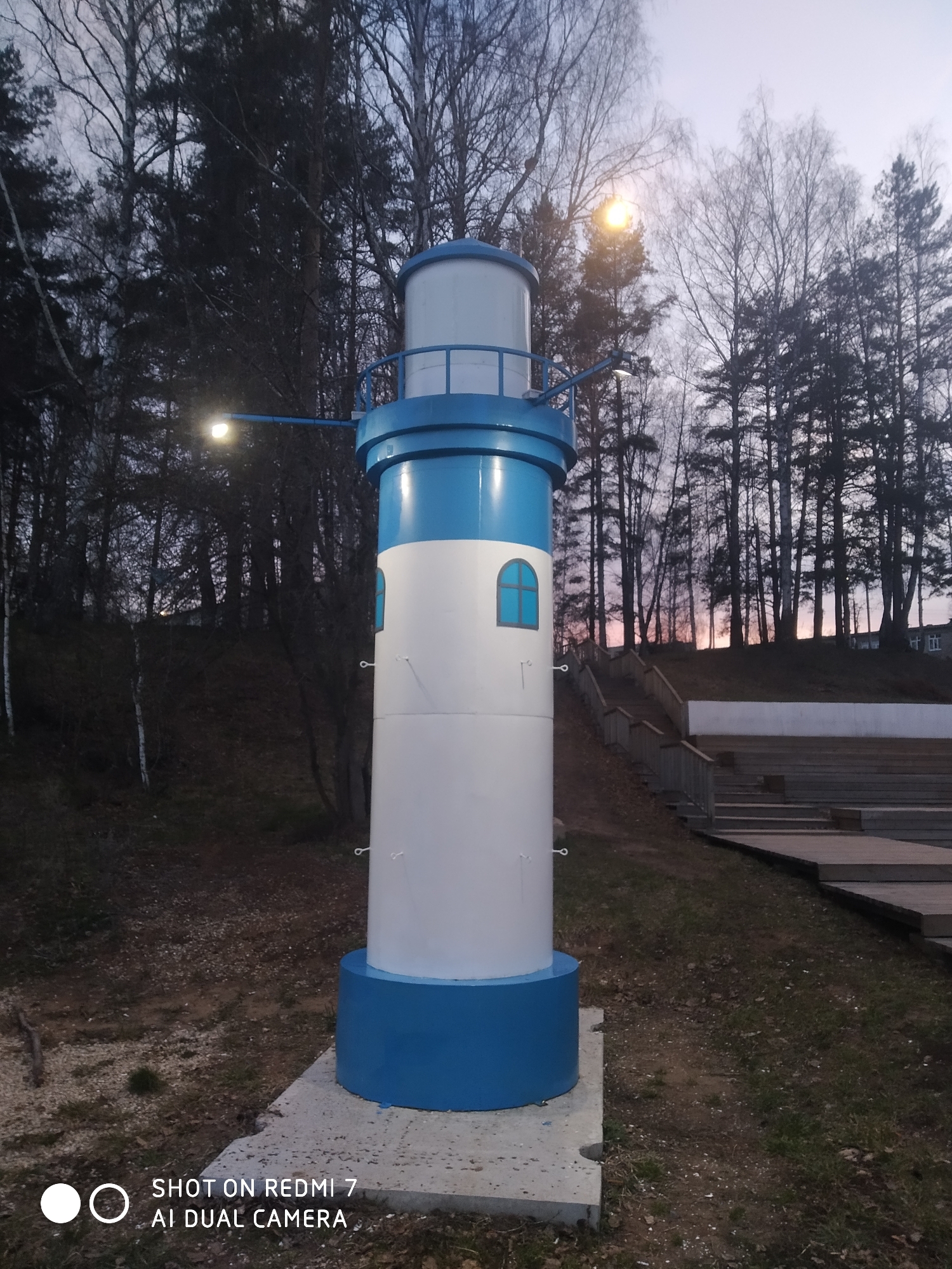 A vertical shot captures a distinctive, decorative lighthouse-like structure, painted in white and blue, standing prominently in an outdoor setting in Chaikovsky, Russia. The cylindrical tower features a broad blue base, a white body with two small arched blue windows, a blue mid-section with a railing, and a white top. Two streetlights extend from the blue band, with the one on the left clearly visible and illuminated, casting a bright glow.
Behind the lighthouse, a dense forest of various trees, including tall pines and bare-branched birches, extends across the background and up a gentle slope. To the right of the lighthouse, wooden stairs ascend the slope, leading to a visible section of tiered wooden seating, suggesting an outdoor amphitheater or public viewing area. The ground around the structure is uneven, covered with dirt, sparse grass, and some dry foliage.
The image appears to be taken during dusk or early morning, evidenced by the dim ambient light and the pale sky, which shows faint hints of pink and orange on the horizon behind the trees. The illuminated streetlights on the lighthouse confirm the low light conditions. No people are present, contributing to a tranquil and still atmosphere. A watermark in the bottom left corner reads "SHOT ON REDMI 7 AI DUAL CAMERA".